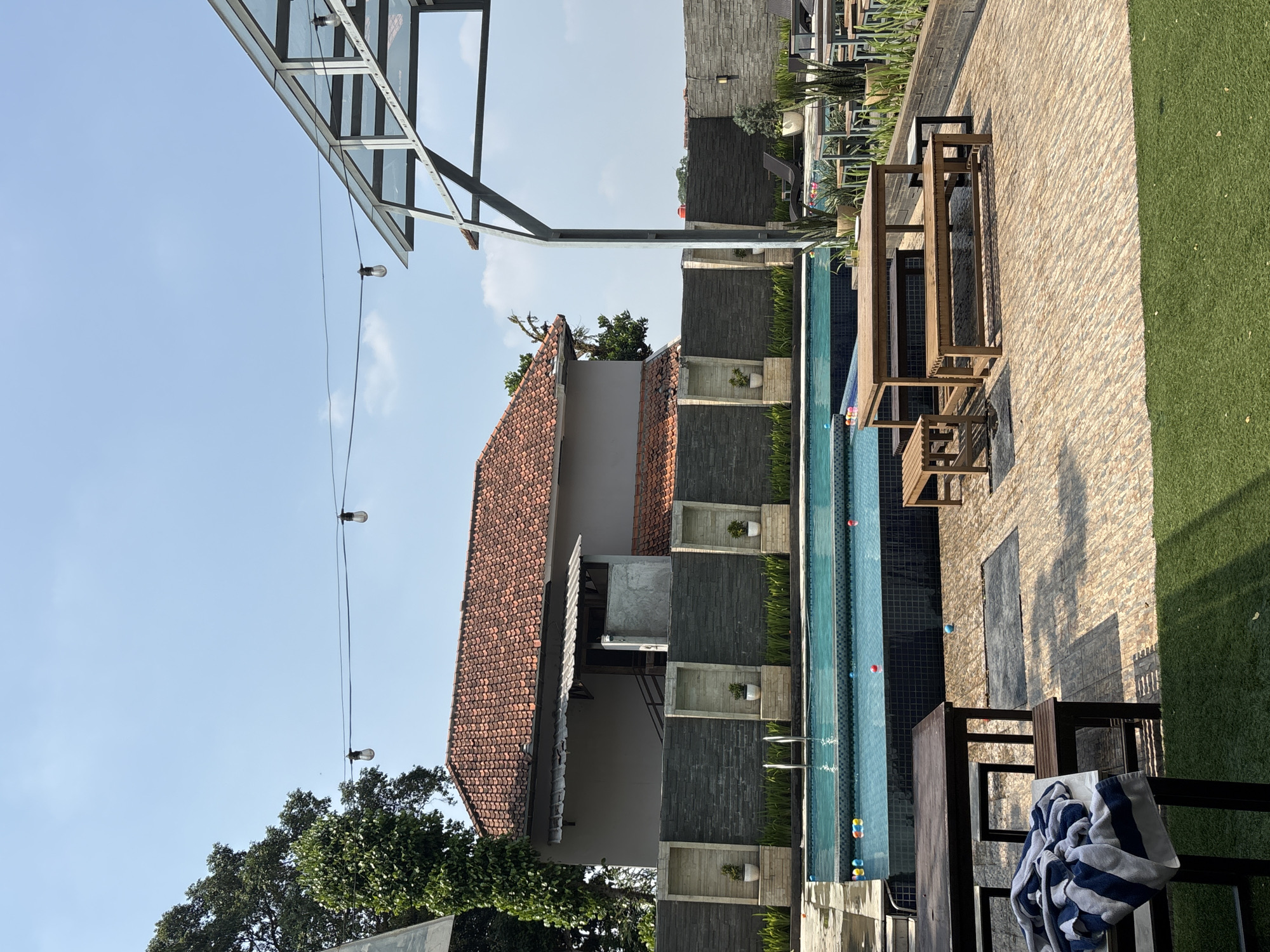 The image shows an outdoor swimming pool area in Binong, Indonesia. In the foreground, there is a wooden table and benches set on a paved patio. A striped towel is draped over one of the benches. Beyond the patio is a swimming pool with steps leading into it, and colorful balls floating on the surface. A dark gray stone wall with decorative niches containing small potted plants borders the pool area. Behind the wall is a multi-story building with a red tiled roof. Overhead, strings of lights are suspended against a clear blue sky. To the right, a modern, open-air structure with a metal frame and glass roof is visible, along with more outdoor seating and potted plants. The overall scene suggests a resort or a venue designed for relaxation and recreation.