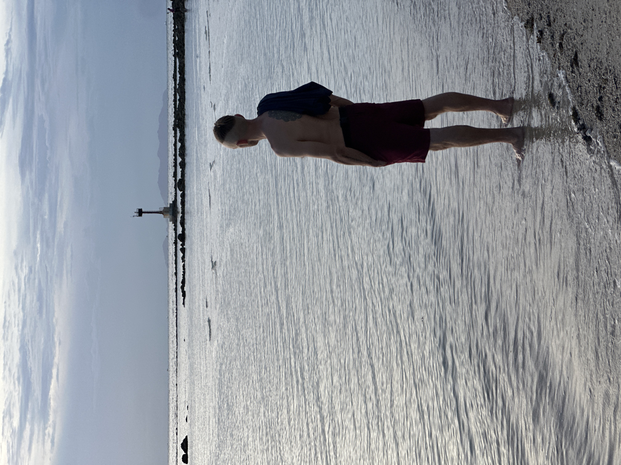 A full-body, outdoor, low-angle shot shows a man from behind, standing at the edge of the water on a beach. The man is facing the sea and has dark hair styled with an undercut and a tattoo on his upper back. He is wearing dark red swim shorts and has a dark navy cloth draped over his right shoulder. The water is shallow, reaching his ankles, and the sand is visible along the shoreline. In the distance, a lighthouse structure is visible on a rocky outcrop, and the sky is cloudy with a soft, diffused light. The scene suggests a serene, possibly late afternoon or early morning, coastal environment. This setting is consistent with the beaches found in ตำบลเกาะพะงัน, Thailand, known for its coastal landscapes and natural beauty.