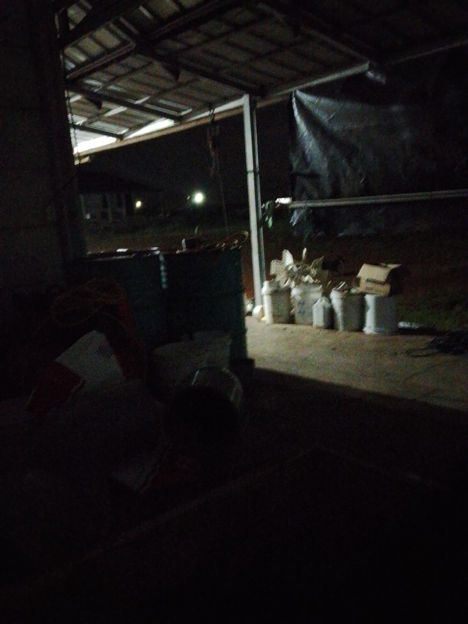 The image shows a dimly lit outdoor storage area, possibly at night, in Bukit Ketok, Indonesia. The area is covered by a partially enclosed structure with a metal roof and support beams. Various objects are present, including large barrels, bags, and containers. Some containers appear to hold plants or refuse. A dark tarp is visible in the background. The overall scene is cluttered and poorly illuminated. In the distance, lights from buildings are seen.