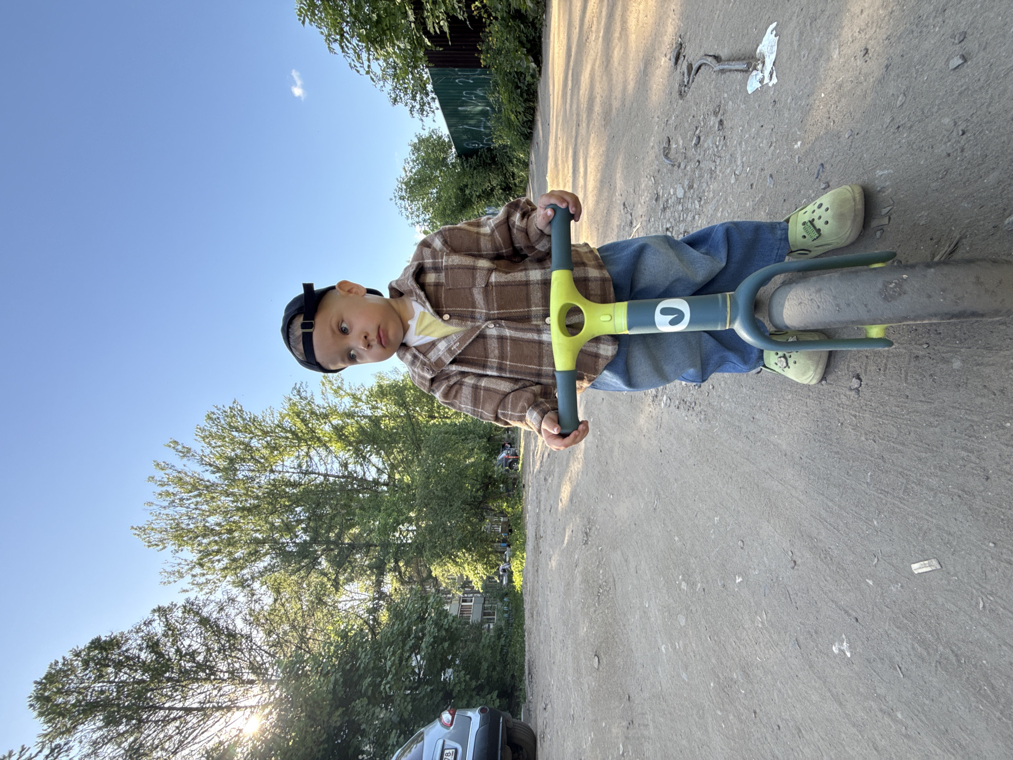 A child in a brown plaid shirt, blue jeans, and light green shoes stands on a gray balance bike on a paved surface. The child wears a dark baseball cap backwards. The bike has a light green handlebar stem and a gray frame. The surrounding environment includes trees, a partial view of a car in the lower left corner, and a fence with some writing on it. The location is likely Rusanovo, Russia, based on prior knowledge and image context.