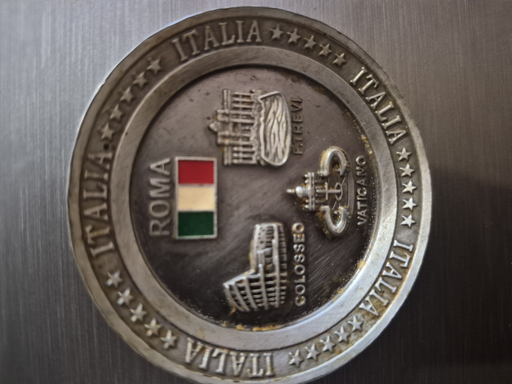 A circular, aged metallic souvenir coin or medal is depicted in a close-up, top-down shot. The object, made of silver-toned metal, exhibits noticeable tarnishing and some yellowish-brown residue accumulated in its crevices. It is resting on a subtly reflective, brushed metallic surface.
The coin's design celebrates Italian landmarks and symbols. Its raised outer rim prominently features the word "ITALIA" repeated four times, with groups of five-pointed stars separating each instance of the word.
The central, slightly recessed area of the coin showcases four distinct elements. On the left side, a miniature Italian flag with vertical green, white, and red stripes is depicted, accompanied by the vertically inscribed text "ROMA". Above the flag and to the right, an architectural rendering of the Trevi Fountain is shown, with the vertical text "F.TREVI" (likely short for Fontana di Trevi) beside it. Below the fountain, symbols associated with the Vatican, including a papal tiara and crossed keys, are illustrated, alongside the vertical word "VATICANO". Completing the central design, below the Vatican symbols and to the left, is a representation of the Colosseum, with "COLOSSEO" inscribed vertically next to it.
The object appears to be a commemorative piece, possibly a magnet or similar souvenir, highlighting iconic aspects of Rome and Italy. There are no people visible, and the lighting suggests an indoor setting.