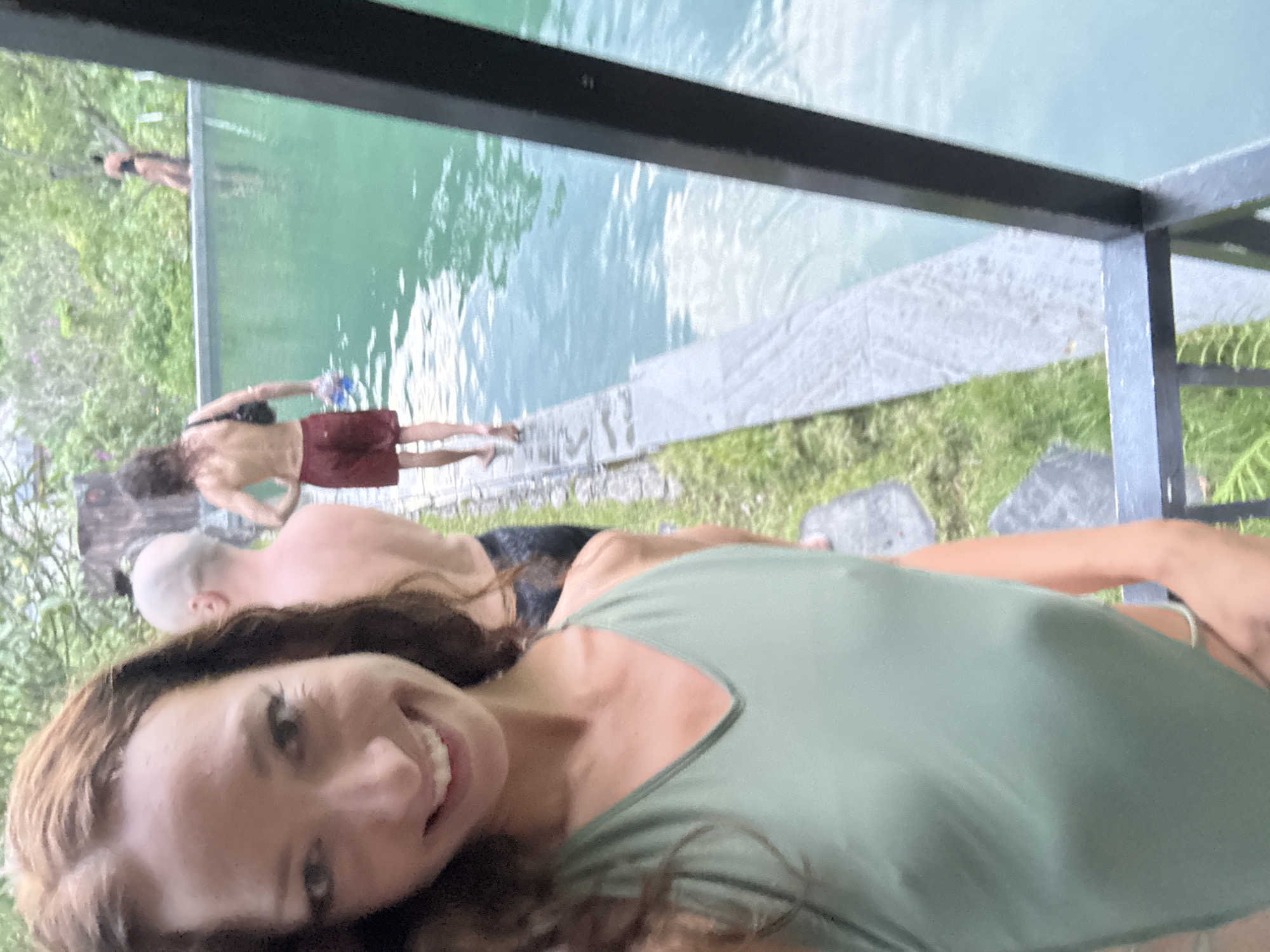 The image, taken in Đà Nẵng, Vietnam, captures a cheerful selfie with a resort-like scene in the background. In the immediate foreground, a woman with long, wavy brown hair and a light green top smiles brightly at the camera, holding the phone to take a selfie. Her expression conveys happiness and relaxation.
Behind her, the scene unfolds around a swimming pool with clear, turquoise water. A dark metal frame and a glass railing separate the selfie-taker's vantage point from the pool area. Several other individuals are visible:
One person with a bald head is partially visible to the left of the smiling woman, seemingly relaxing on the ground or a deck by the pool.
Further back, a person with long, dark, curly hair, wearing red shorts or swim trunks, stands at the edge of the pool, facing away from the camera, holding a blue item in their hand, possibly about to enter or exit the water.
In the background, a few other figures are indistinctly visible in or around the pool, partially obscured by the glass barrier and surrounding greenery.
Lush green foliage and trees line the perimeter of the pool area, suggesting a natural or landscaped setting. The bright, even lighting indicates it is daytime, likely a sunny and pleasant day. The overall atmosphere is leisurely and joyful, indicative of a vacation or relaxed gathering. No visible text is present in the image.