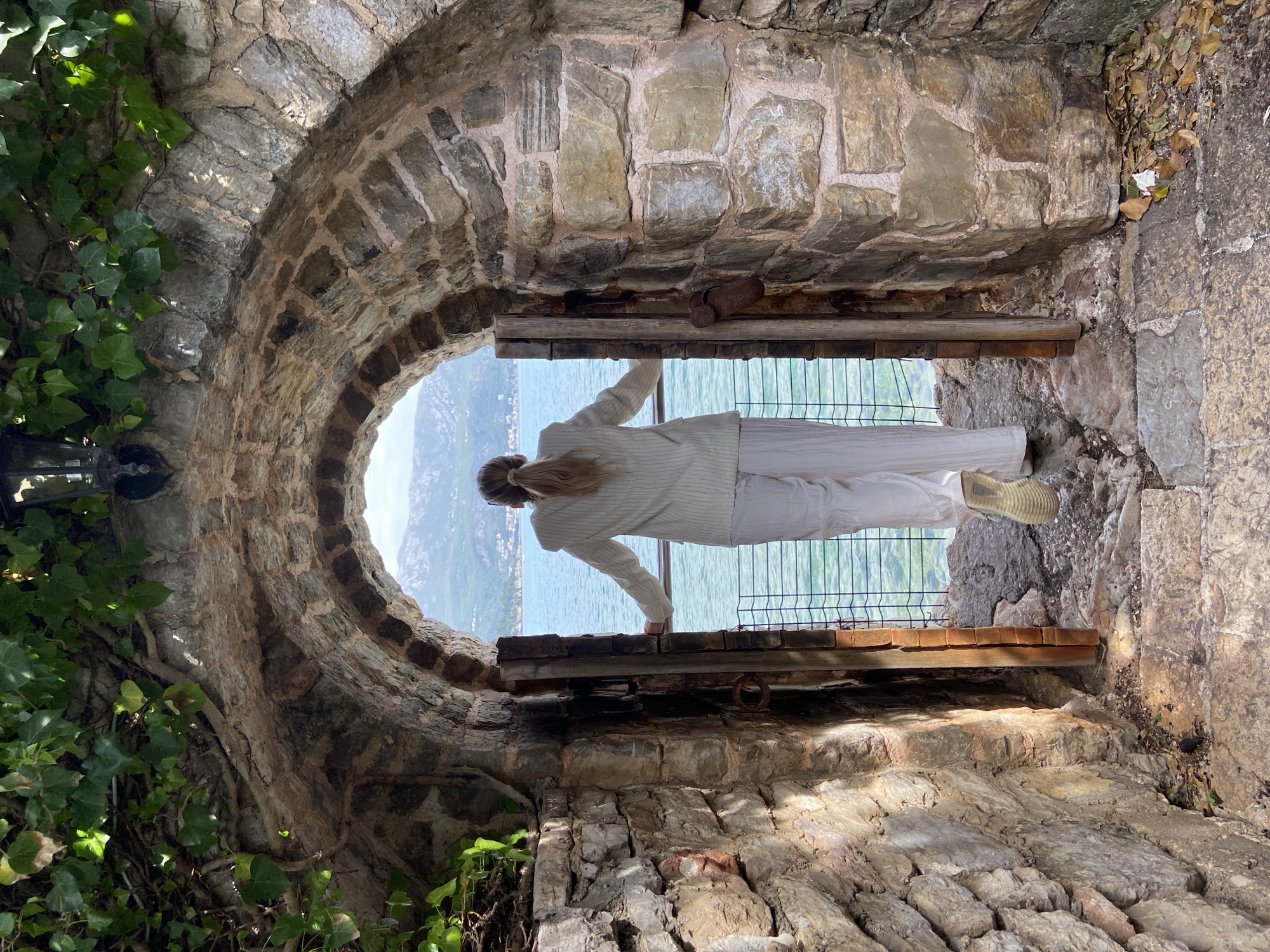 A person is seen from behind, standing in a rustic stone archway and looking out over a scenic vista. The image is oriented vertically, with the archway forming a prominent, dark frame. The archway is constructed from irregular, rough-hewn stone blocks, with green ivy clinging to the stone on the left side.
The person, a woman, wears light-colored clothing, consisting of a striped long-sleeved top or jacket and trousers. Her hair is tied back. She is leaning on a wooden sill or ledge within the archway, with a dark metal mesh fence or railing visible below her hands.
Beyond the archway, an expansive view unfolds, featuring a large body of water, likely the sea, which appears calm and blue-green. In the distance, mountains or hills rise from the water's edge, partially obscured by a hazy sky. Sunlight illuminates parts of the stone structure, casting shadows, indicating it is daytime. The floor within the stone structure is also made of similar stone pavers, with some dry leaves visible on the right side.
The scene depicts a peaceful moment of contemplation, with the ancient stone structure providing a dramatic frame for the natural beauty of the coastal landscape, consistent with a location like Budva, Montenegro. There is no visible text in the image.