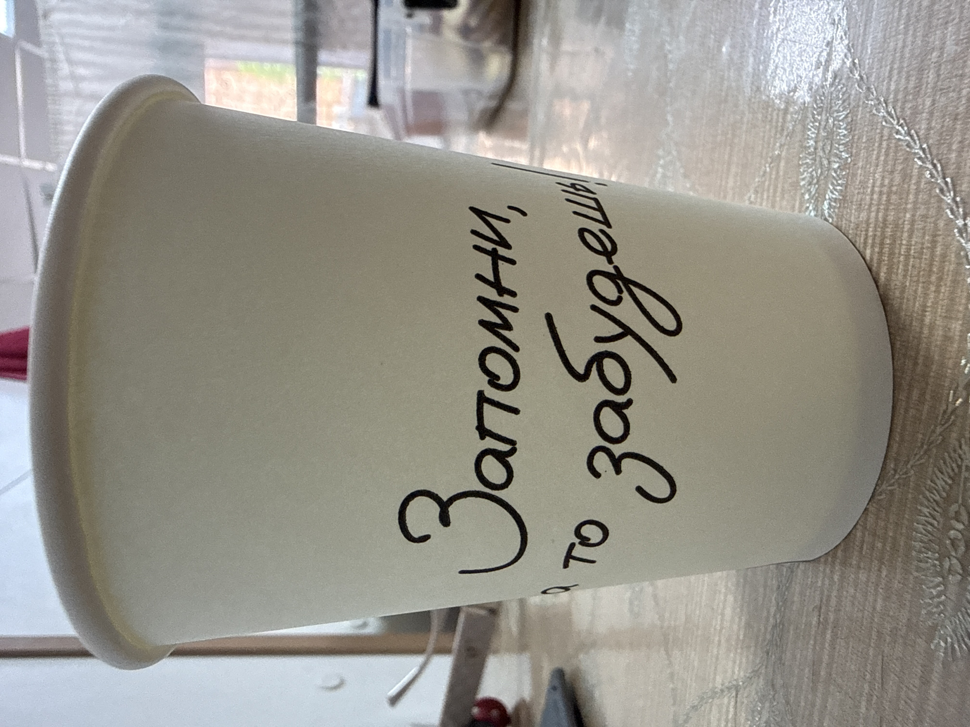A light-colored paper cup sits on a table. The cup has black handwritten text on it in Russian. The text reads "Запомни, а то забудешь!" which translates to "Remember, or you will forget!". The cup is positioned centrally in the frame, with the background slightly out of focus. The background suggests an indoor setting, possibly a home or cafe, with a window and blinds visible. The table appears to be a wooden surface with a patterned cloth underneath. There are no people visible in the image. The location is identified as Beslan, Russia, though no specific landmarks or context within the city are provided beyond that.