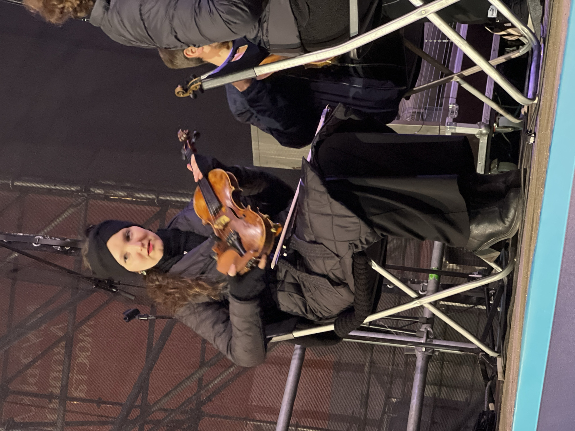 In an outdoor setting at night, a woman wearing a black beanie, dark winter coat, and black gloves is seated on a metal folding chair and playing a violin. Another person is seated behind her, also playing a violin. Both are positioned on a metal scaffolding structure against a backdrop of red and grey material with faint text, which may indicate the Saint Petersburg location. The scaffolding and a turquoise panel indicate the presence of a stage or temporary structure.