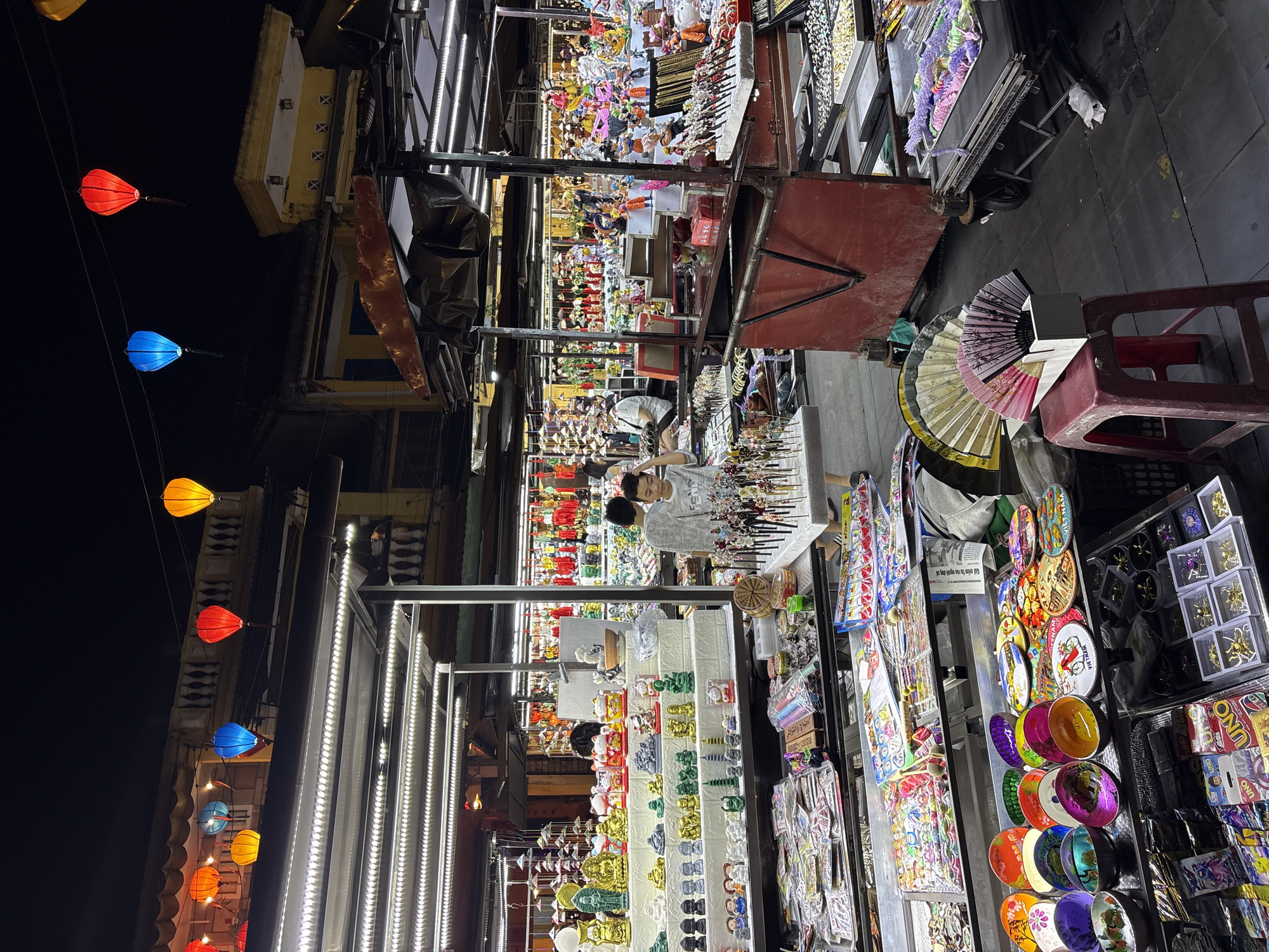This photograph shows a night market scene in Hội An Ward, Vietnam. Lanterns in blue, orange, and red are hung overhead. A narrow walkway is flanked by vendor stalls displaying a variety of goods including souvenirs, jewelry, ornaments, and fans. People, including a couple, are browsing the items on display. The stalls are brightly lit, contrasting with the darker sky above. A red plastic stool is positioned near one of the stalls. The ground is paved and slightly damp.