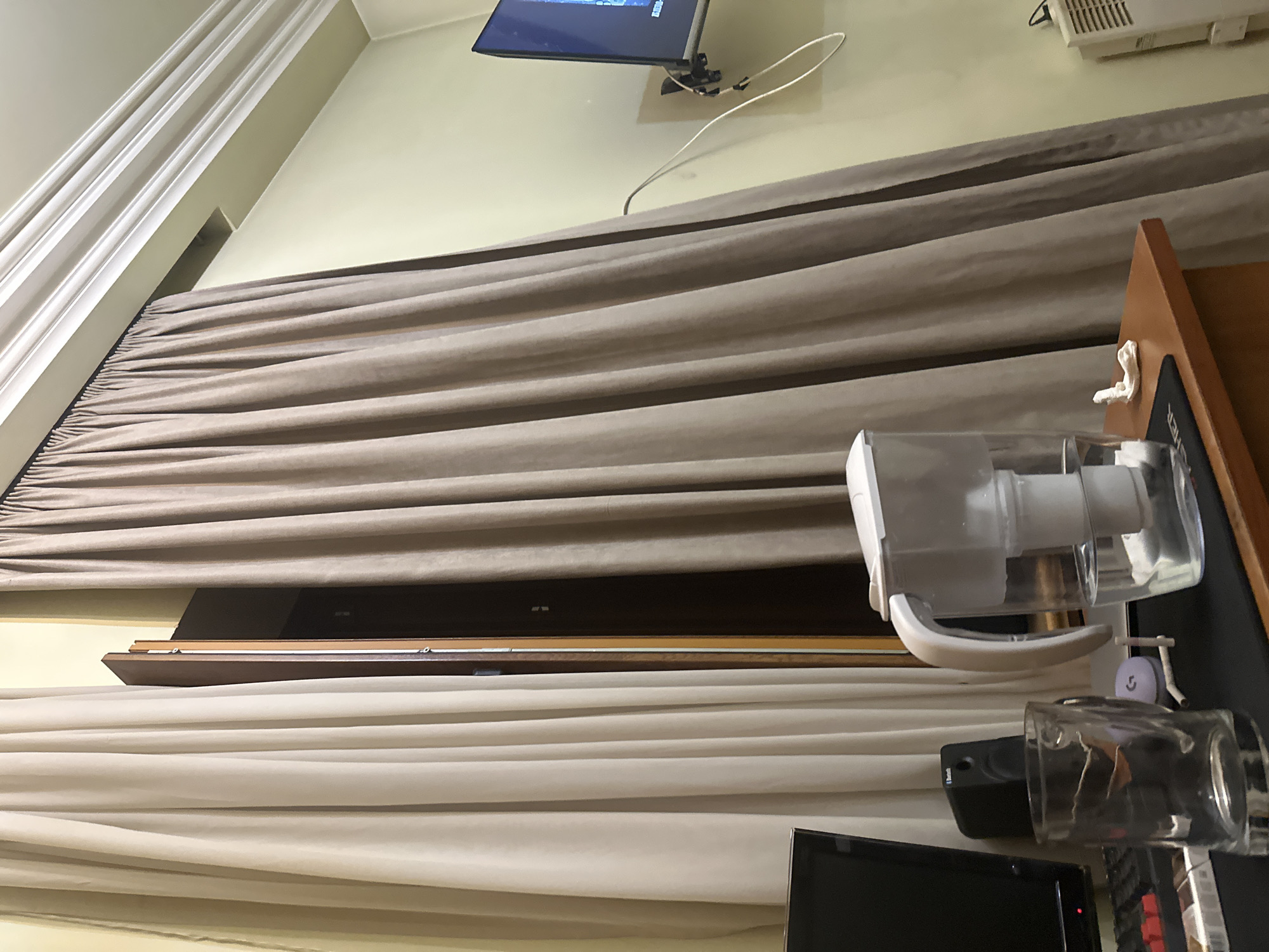 The image depicts an indoor scene, possibly a home or office setting, in Unknown, Unknown. The angle is unusual, seemingly taken from a low perspective looking upwards. The most prominent feature is the layered curtains covering a window or possibly a doorway. The top layer is a light brown or beige color, while the bottom layer appears to be a lighter shade, possibly white or cream. The curtains are heavily draped and create a strong horizontal emphasis across the image.
A television is mounted on the wall in the upper portion of the frame, with visible cords extending from it. Near the right edge, a desk or table is visible, with a water filter pitcher and a glass sitting on it. Parts of a computer monitor and keyboard can also be seen on the table.
The color palette is neutral, dominated by shades of beige, brown, white, and black. The lighting appears to be artificial, casting soft shadows on the surfaces. There are no people visible in the image.