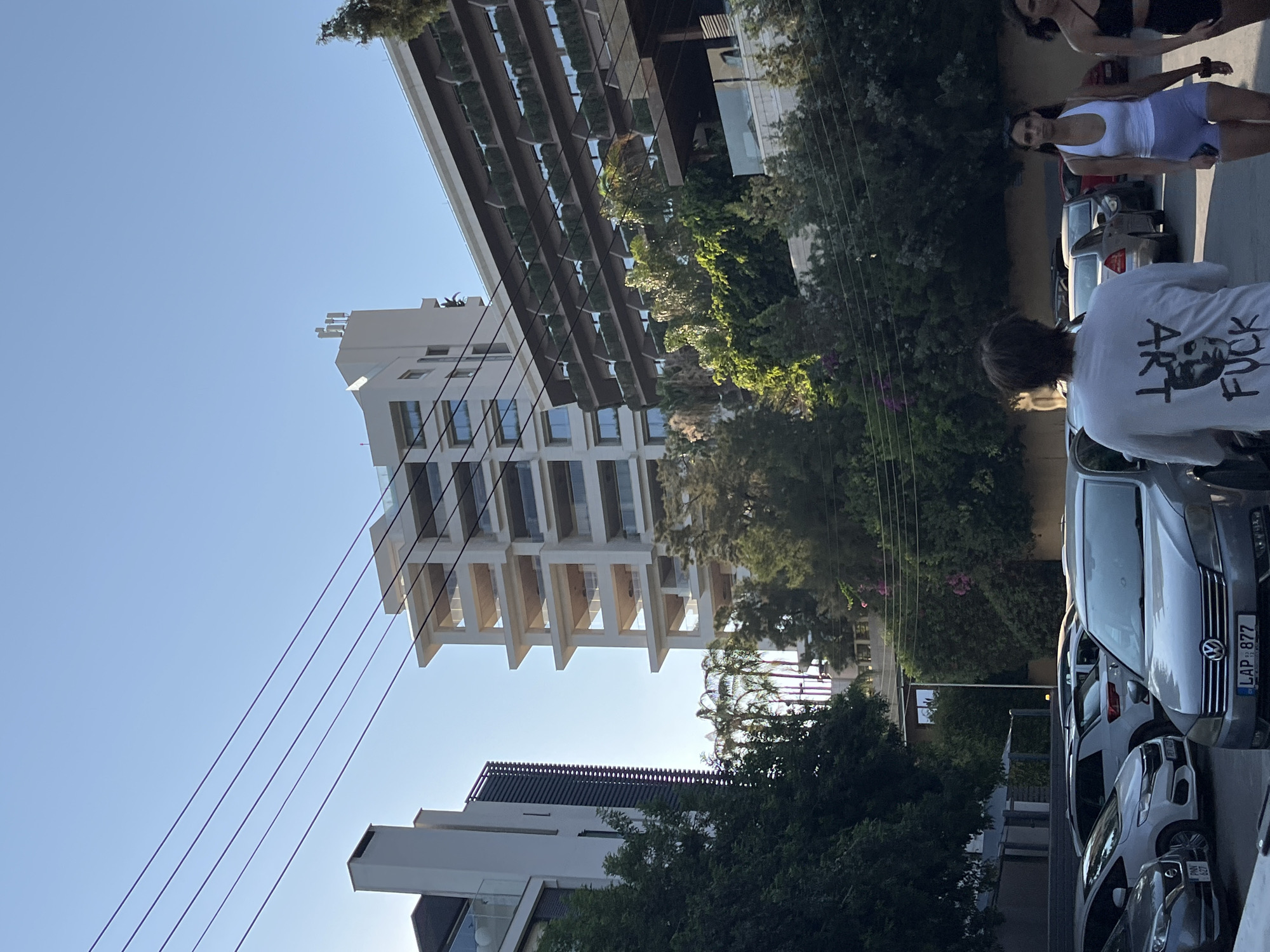 In Agios Tychona Community, Cyprus, an outdoor scene captures a bright, clear sky with several parallel power lines stretching across the upper portion of the frame. A modern, multi-story white building with a stepped facade and balconies is prominent on the right. To its left, another tall, white building with a more angular design stands, featuring vertical slatted elements. In the foreground, a parking lot is filled with several cars, including a silver Volkswagen Passat with license plate LAP 877. Three individuals are walking away from the camera. The person in the foreground wears a white t-shirt with graphics on the back, the central figure is a woman in a white tank top and light purple shorts, and a third woman in a black sports bra and black shorts is to her right. Lush green trees and foliage surround the buildings and parking area. The lighting suggests it is daytime, likely late afternoon due to the long shadows cast by the trees.
