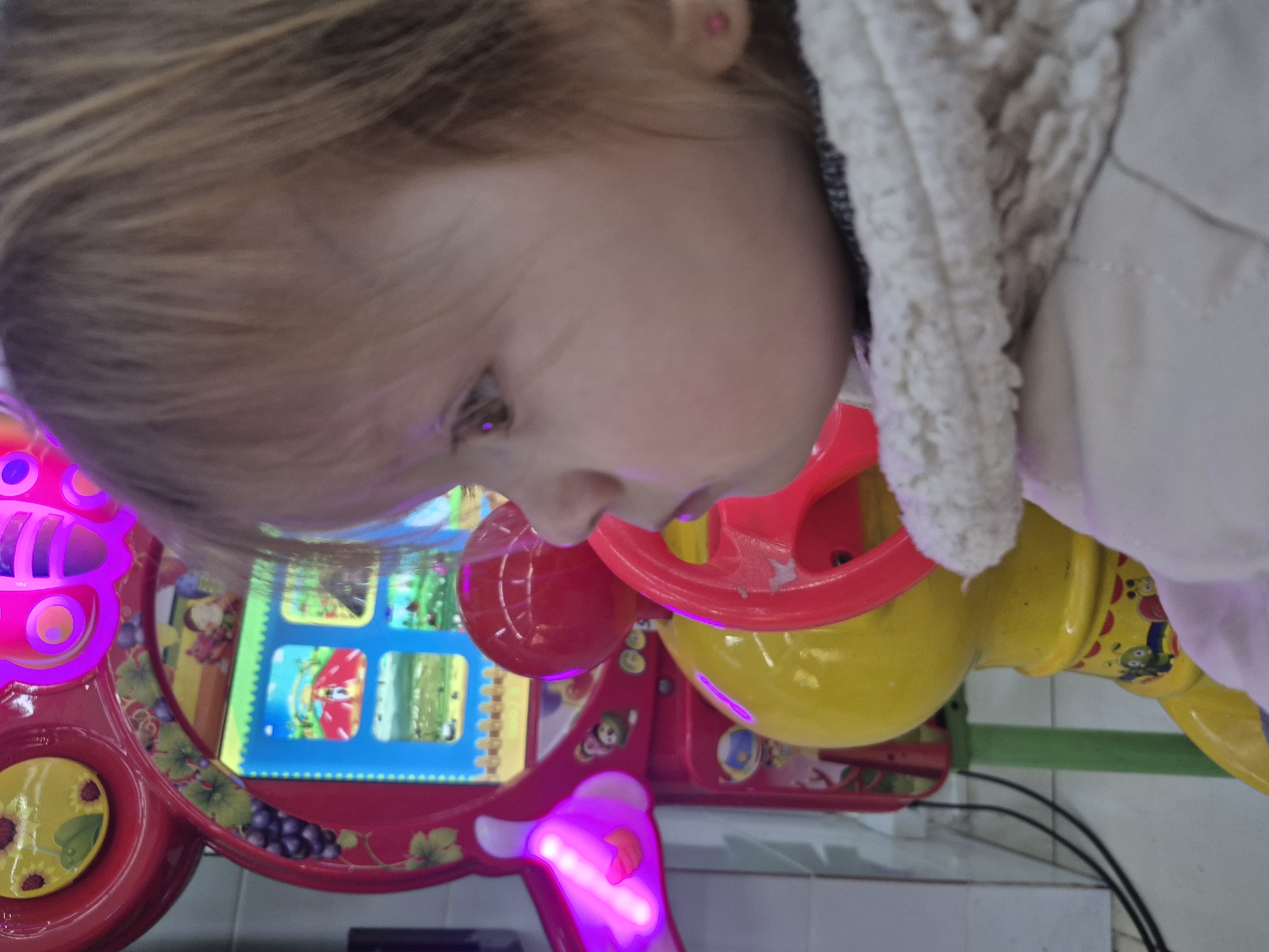 A toddler with blonde hair is looking at a brightly colored, interactive toy or game. The toy has a screen with various images displayed, and it features components in red, yellow, and pink with decorative patterns. The child is wearing a light-colored jacket. The background appears to be a commercial or public space with tiled flooring.