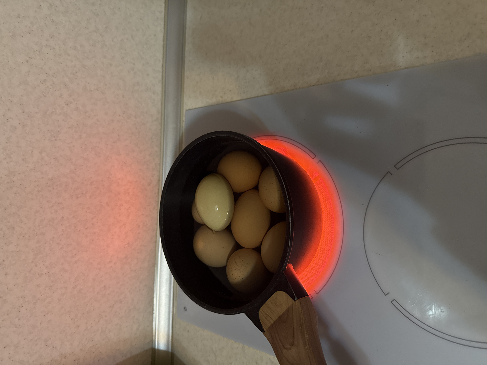 A top-down shot depicts a small, black saucepan filled with approximately eight hard-boiled eggs, sitting on a white electric stovetop. The burner beneath the pan is glowing red, indicating it is in use. The pan has a wooden handle. The stovetop is clean and features circular markings delineating the heating element. The background includes a light-colored wall with a slightly textured surface. There is no evidence to confirm this photo was taken in Varna, Bulgaria.