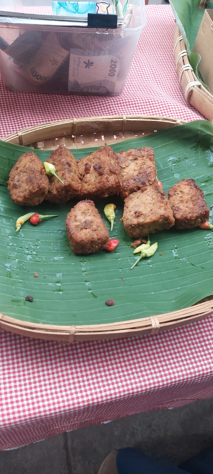 A close-up, top-down shot shows a serving of fried tempeh, cut into bite-sized pieces, arranged on a large green banana leaf. The tempeh pieces are irregular in shape and appear to be coated in a light batter. Several small red and green chilies are scattered amongst the tempeh. The banana leaf is resting on a woven bamboo tray, which is itself placed on a red and white checkered tablecloth. In the background, a clear plastic container is visible, filled with Indonesian currency notes, predominantly in denominations of 2000 and 10000. A black binder clip is securing the lid of the container. The overall setting suggests a street food stall or a casual market stall in Lumajang, Indonesia, possibly a roadside vendor where one might purchase food and potentially exchange money or conduct small transactions. The image is shot with a shallow depth of field, blurring the background and focusing on the food.