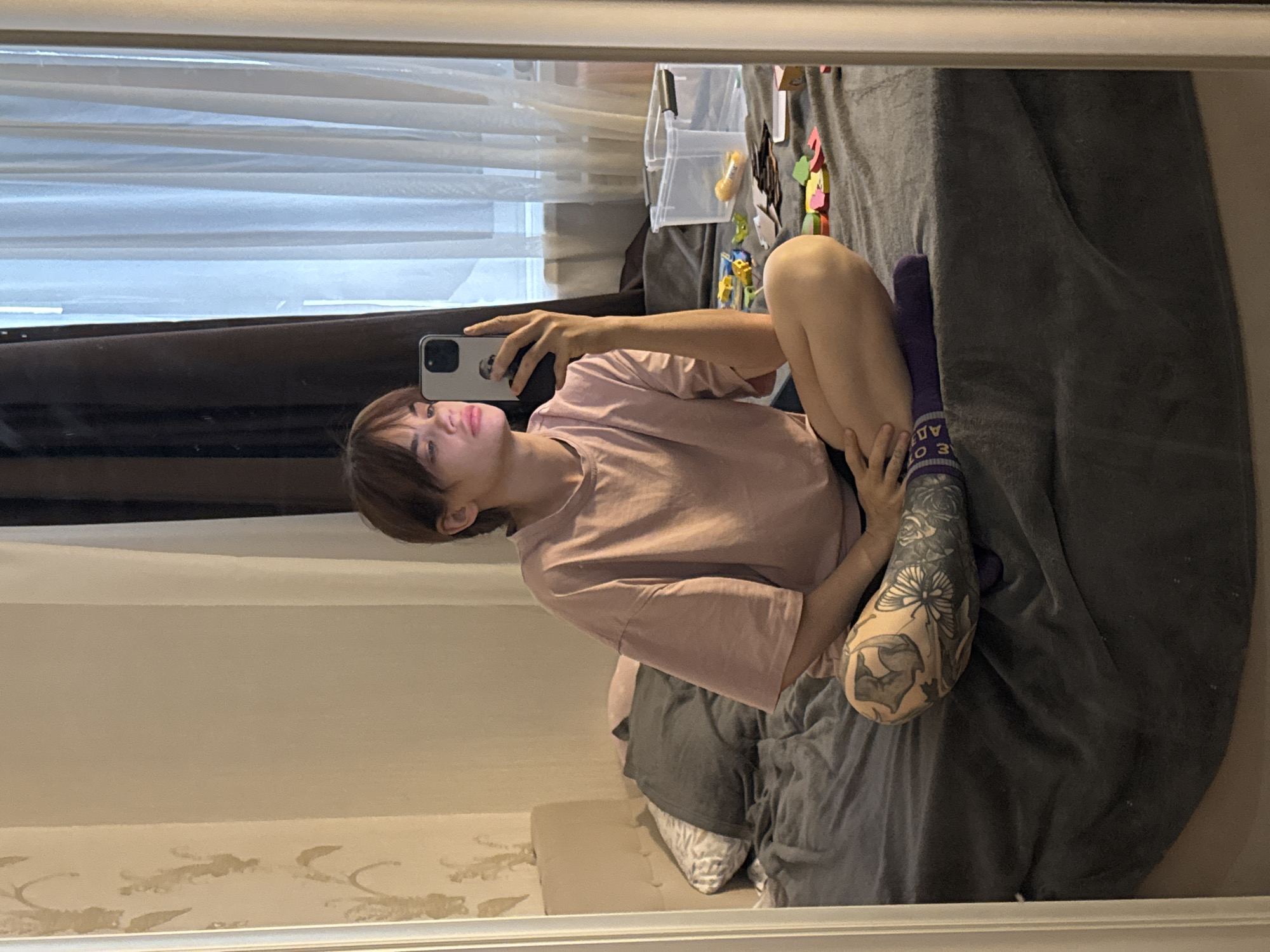 A young person with short brown hair, wearing a pink t-shirt and purple socks with white lettering, is taking a selfie in a mirror. The person is sitting on a bed covered with a gray blanket. The left arm is prominently displayed, featuring a large tattoo that includes roses and a butterfly. The background includes a window with sheer white curtains and a darker brown curtain partially obscuring it. To the right of the bed, there is a clear plastic container with miscellaneous items inside, including colorful objects. The room's wall is visible at the bottom of the mirror, showing wallpaper with a subtle pattern. There is no explicit indication of the city of Rostov-on-Don, Russia, in the image.