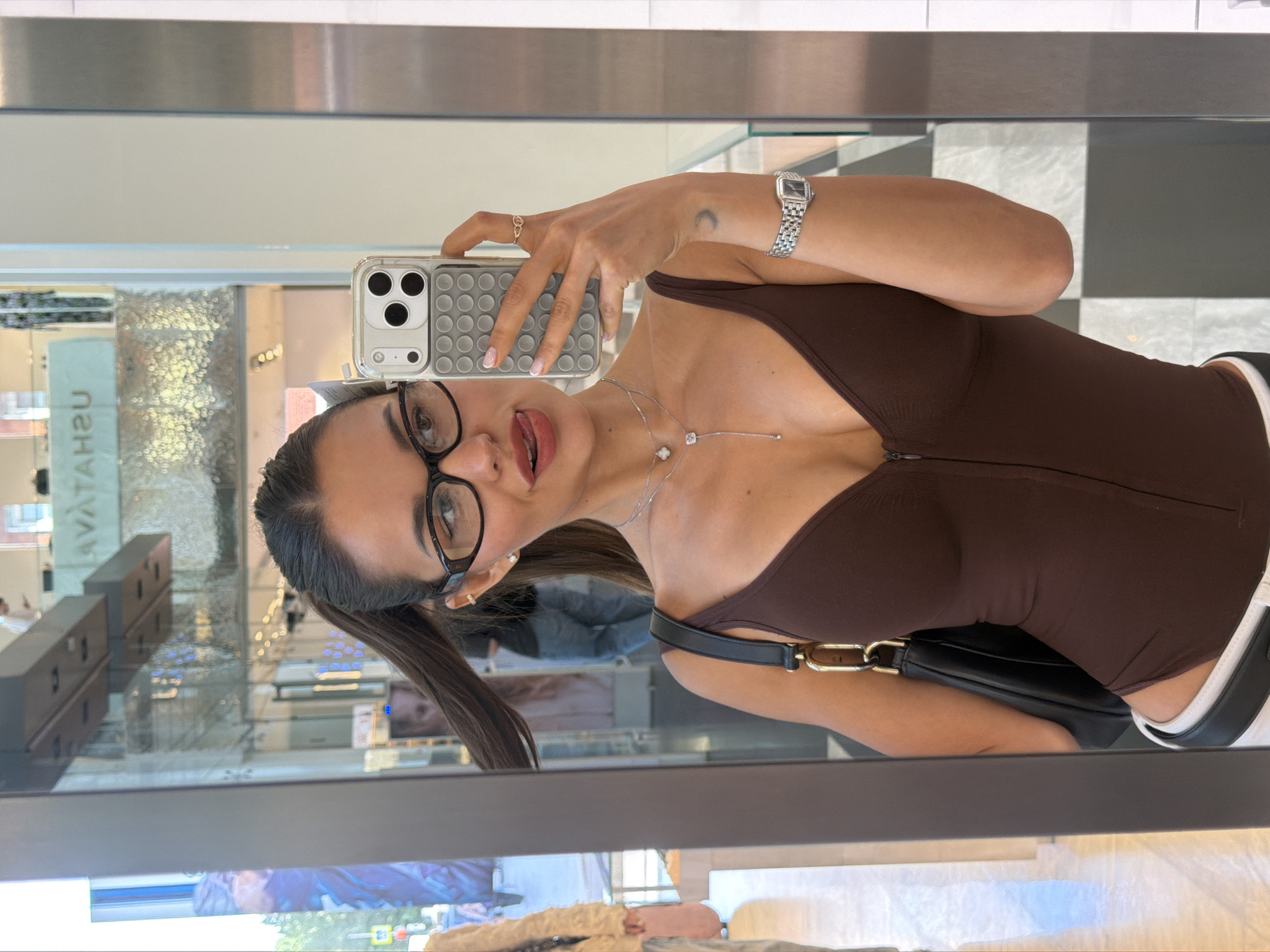 The image is a mirror selfie featuring a young woman captured from the chest up, seemingly taken indoors. She holds a smartphone with a clear case and a gray, textured pop-it style accessory on the back, covering part of her face. Her dark hair is pulled back into ponytails, and she wears black-rimmed glasses. Her expression is playful, with her tongue slightly extended.
She is adorned with a silver-toned watch on her left wrist, a subtle gold-toned ring on her right ring finger, and a delicate silver necklace featuring two charms—one resembling a clover and the other a small, square-cut stone. She also has small stud earrings. Her attire consists of a form-fitting, dark brown top with a front zipper detail. A black shoulder bag with gold chain-link accents is slung over her shoulder. Hints of white and black clothing or accessories are visible near her lower right arm. A small crescent moon tattoo is visible on her left forearm.
The background, visible as a reflection in the mirror, suggests an indoor commercial or public space, possibly a modern shopping center or large store. Reflections show elements of interior architecture, glass partitions, and display areas. A black and white checkerboard pattern is visible on a surface behind her right arm. Text reflected on a sign in the background reads "UATAVA" (which would be "AVATAN" if unreflected), possibly indicating a store name. The overall lighting appears artificial, characteristic of an indoor commercial environment. The scene is an indoor location, potentially a shopping center in Moscow, Russia, as per the provided context.