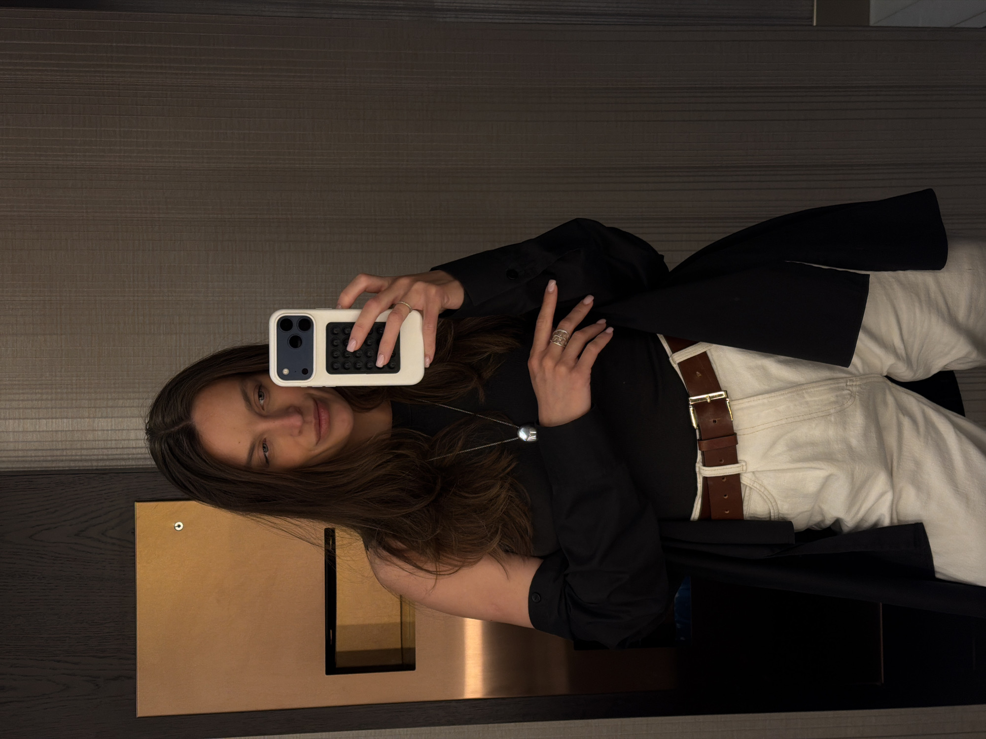 A young woman with long brown hair takes a selfie in a mirror. She is wearing a black shirt, white pants with a brown belt, and a black jacket. She holds a white smartphone in her left hand. The background consists of a beige wall with a dark gray horizontal pattern, and a mirrored surface with a gold-colored panel. The scene appears to be indoors, likely in a hotel room or similar setting. It is not possible to determine the location in the city of Budva, Montenegro based on the image provided.