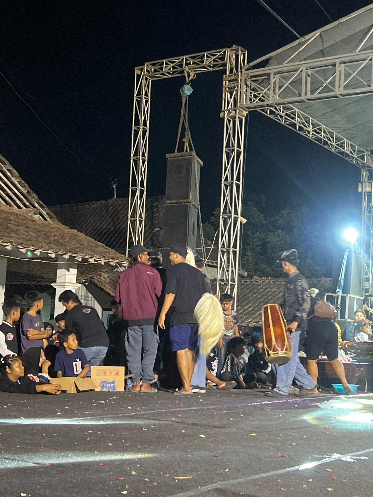 The image depicts an outdoor nighttime scene in Madyocondro, Indonesia, likely at a local event. A stage structure, constructed from metal trussing, is visible, supporting large speakers and a canopy. A group of people, comprising adults and children, are gathered on the ground in front of the stage. Some individuals are standing, engaged in conversation, while others are seated or lying down.
In the foreground, a woman is lying on the ground, propped up on her elbows, looking towards the left. Next to her are two cardboard boxes. Several children are clustered together, some looking towards the stage, others in different directions. Two men are standing and talking, one wearing a maroon jacket and the other a black t-shirt. Another man, dressed in a black patterned shirt and a cap, holds a large, wooden drum with a reddish-brown skin. He is walking towards the right of the frame.
Behind the people, a traditional building with a tiled roof and white pillars is visible. The ground surface is dark and appears to be paved, with scattered pink and red petals. Lighting from the stage illuminates parts of the ground and the gathering crowd. The background consists of dark trees and the night sky. The overall atmosphere suggests a community gathering or performance.