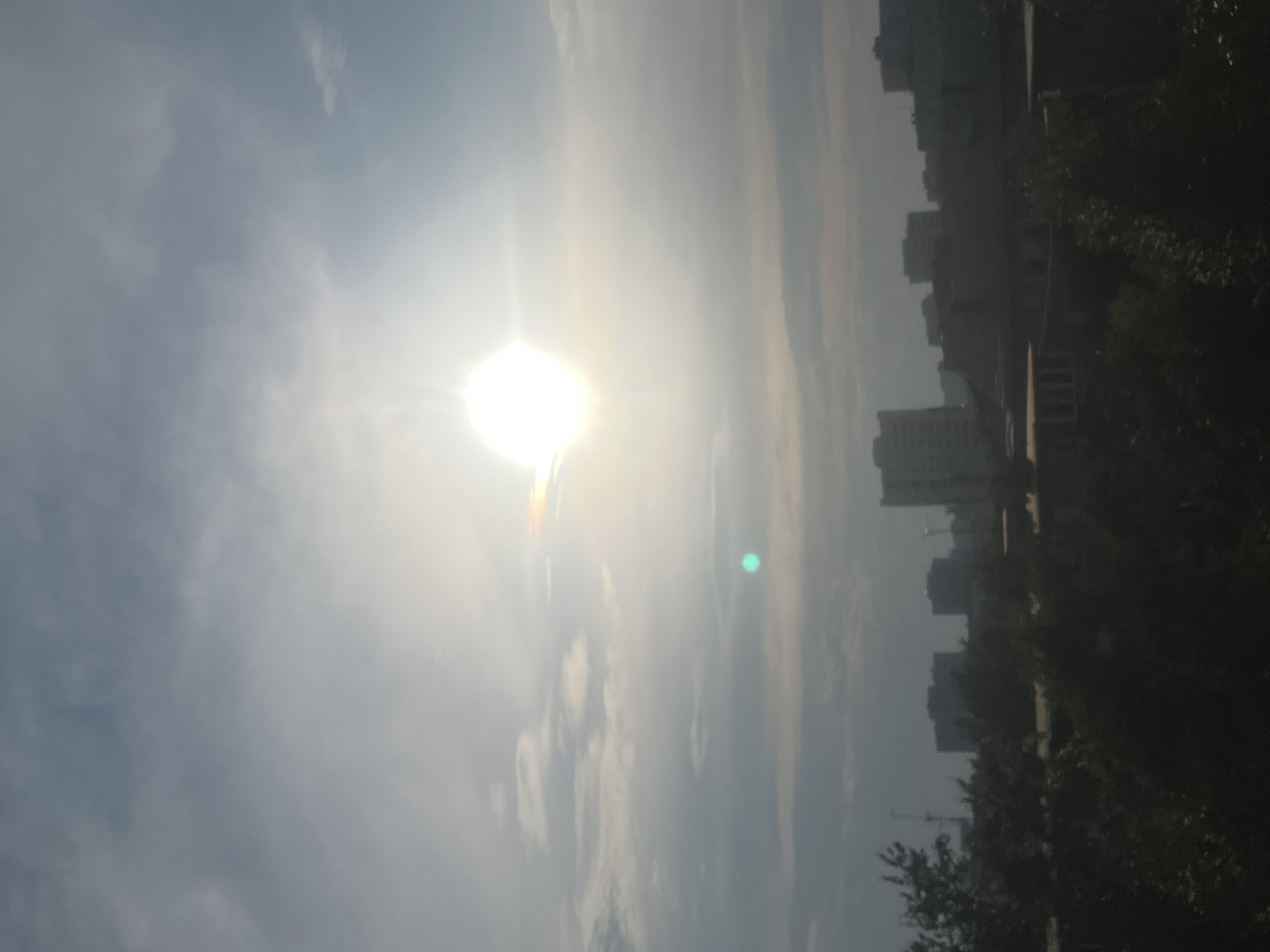 The image depicts an outdoor scene with a bright sun low in the sky, suggesting either sunrise or sunset. The sun is partially obscured by clouds, casting a bright glare across the frame. In the background, a cityscape with multi-story buildings is visible, interspersed with trees. The overall impression is of a somewhat hazy or overcast day in an urban environment. There are no people or distinct objects clearly discernible in the foreground. The presence of buildings and trees suggests a populated area, and the mention of Chelyabinsk, Russia, places the scene within that specific geographical context. The light conditions, with the sun low and the glare, indicate a time of day when light levels are not at their peak. The weather appears to be partly cloudy or hazy, contributing to the soft, diffused light. No text is visible in the image.