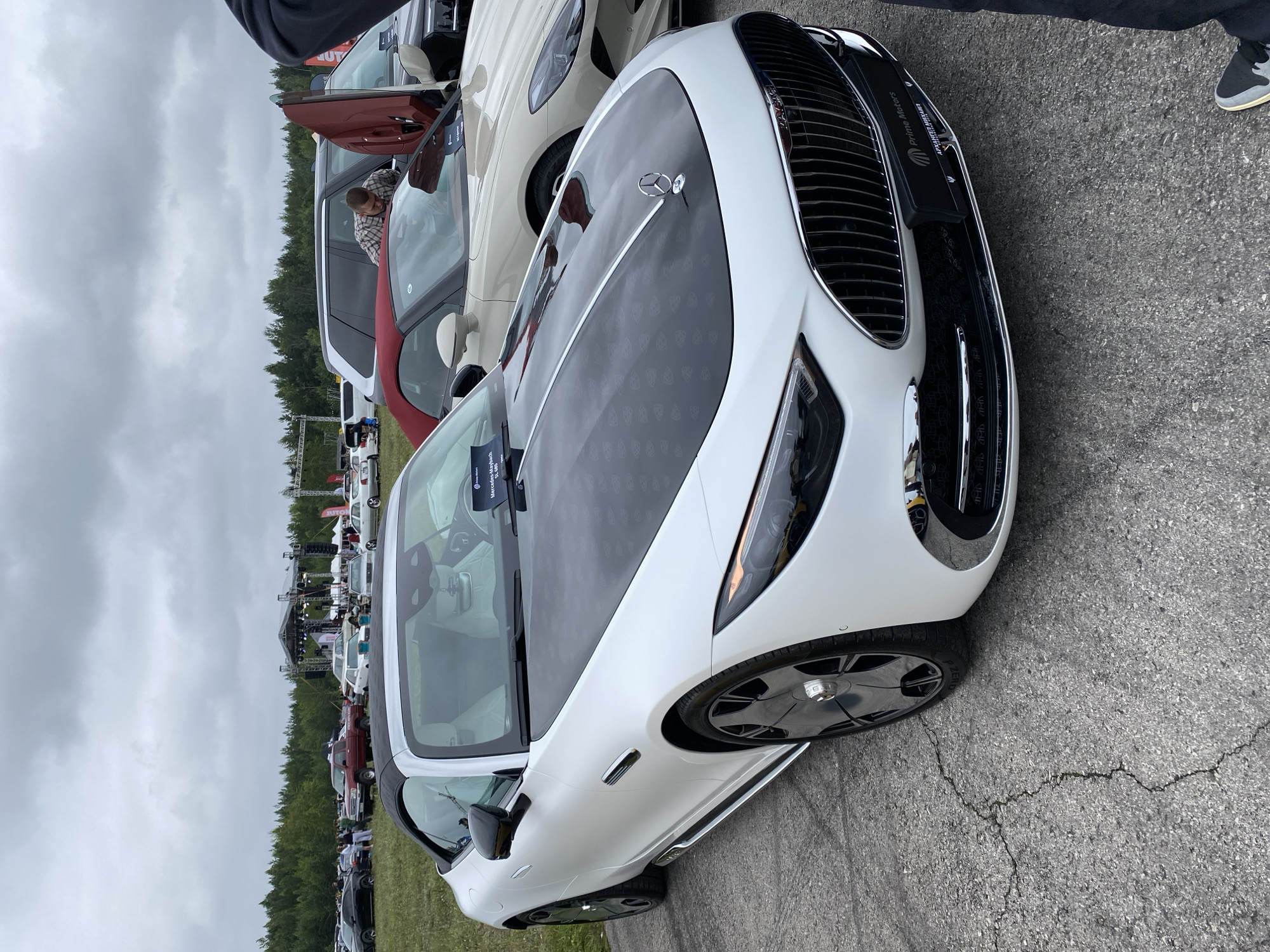 The image shows a white Mercedes-Maybach SL 680 convertible parked on asphalt. The car has a black hood with a patterned design and a chrome grille. A license plate frame from "Prime Motors" is visible on the front of the car. In the background, there is a stage with lighting equipment, suggesting an outdoor event or concert. Several other cars and people are present in the background. The sky is overcast. The location is an open outdoor space, likely a parking lot or field, consistent with an event venue. The provided crops indicate a city setting, but a specific location in Perm, Russia, cannot be determined solely from the visual information.
