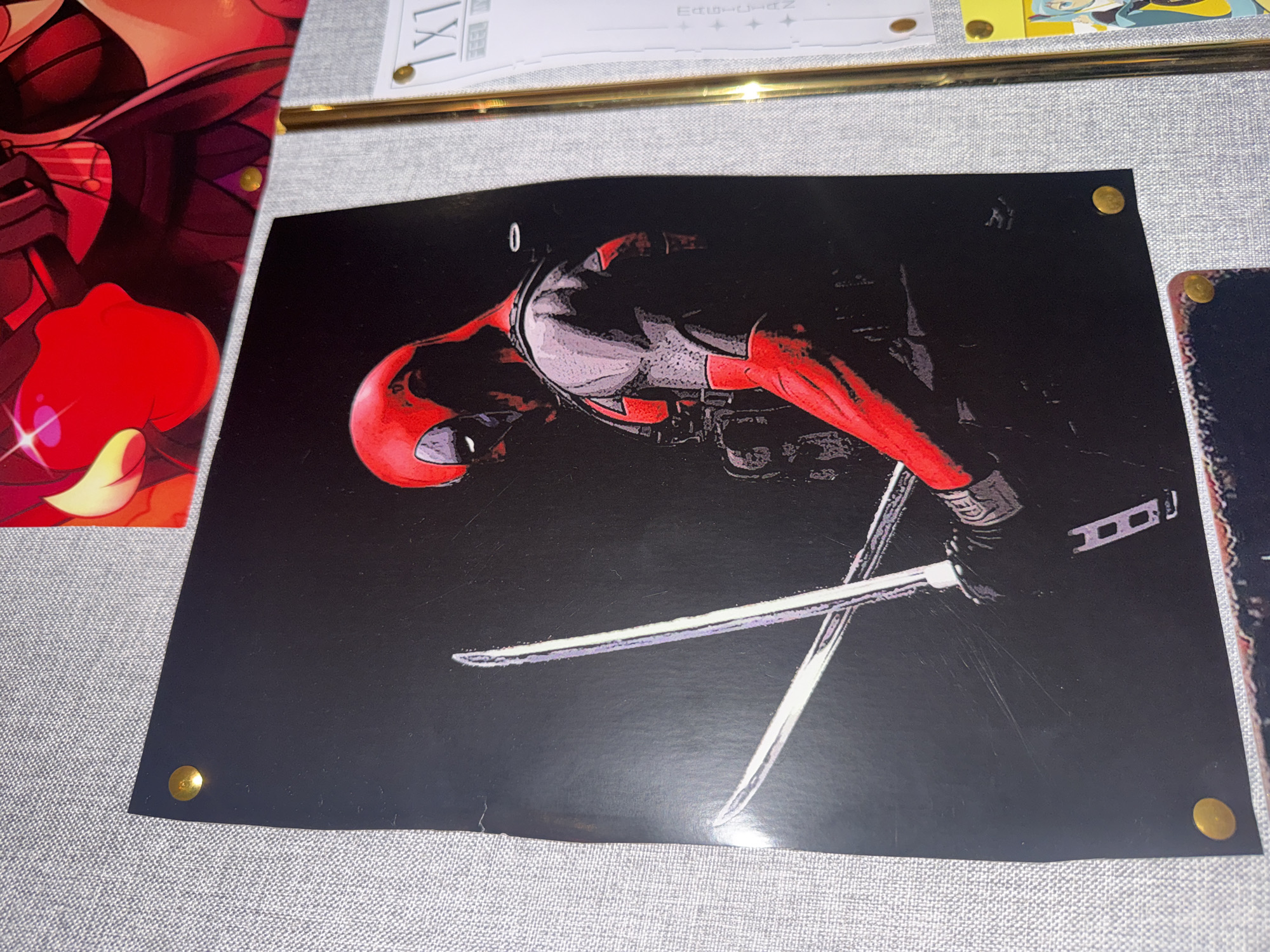 The image shows a close-up of a wall covered in a light gray textured fabric, possibly a bulletin board or a soundproofing material. The wall is adorned with several pieces of art and paper. Dominating the center of the frame is a large, dark poster featuring a stylized illustration of Deadpool. He is depicted in profile, facing left, with his iconic red and black costume. He is holding two katana swords, which are crossed in front of him and appear to be glowing or reflecting light. The background of the poster is solid black.
The poster is secured to the wall with four brass pushpins, one in each corner. To the top of the Deadpool poster, a portion of another artwork is visible, featuring vibrant red and yellow colors, possibly an anime or manga illustration. To the right of the Deadpool poster, a golden frame encloses a white document with text and some symbols. This document is also attached to the wall with a brass pushpin. Below this, another smaller artwork, this one featuring a female anime character with blue hair, is partially visible. This too is secured with a pushpin. A portion of another poster with a dark background and a decorative border is visible at the bottom of the image, also held by a pushpin.
There is no indication of the city of Odesa, Ukraine, or any other specific location in the image. The scene appears to be an indoor display of artwork.