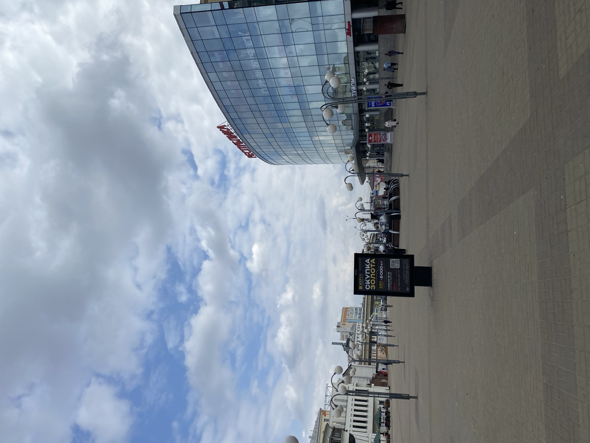The image depicts an urban outdoor scene in Nizhny Novgorod, Russia, featuring a modern building with a reflective glass facade and a paved plaza. The time of day appears to be daytime, under a cloudy sky with some patches of blue.
The primary subject is a large, curved building on the right, clad in blue-tinted glass that reflects the sky and surrounding environment. Visible text on the building includes "туаль" (tual) on a shopfront sign, and reflections of other Russian words like "ФАНТАЗИЯ" (Fantaziya), "ПРИЁМ МЕТАЛЛА" (Priem Metalla - Metal Reception), and "КОМПЬЮТЕРЫ" (Kompyutery - Computers). A red sign on the building's lower section partially reads "СТРОЙМАТЕРИАЛЫ" (Stroymaterialy - Building Materials). Several people are visible walking near the entrance of this building and across the plaza.
The plaza is paved with light-colored blocks. Multiple lampposts with globe-shaped lights line the plaza, extending into the distance. In the foreground, an advertising A-frame sign stands prominently, displaying text in Russian: "АУРУМ СКУПКА ЗОЛОТА" (Aurum Gold Buyout/Purchase). Below this, it details "585 = 8000 р/г" (meaning 585 standard gold purity is purchased for 8000 rubles per gram), a phone number "+7 (831) 266-00-05", a distance indicator "100 м" (100 meters), and "Пл. Революции" (Revolution Square), along with "18+".
The scene suggests a commercial or public area within a city, with pedestrians moving about on a seemingly ordinary day. The weather is overcast but bright, with significant cloud cover dominating the upper portion of the frame.