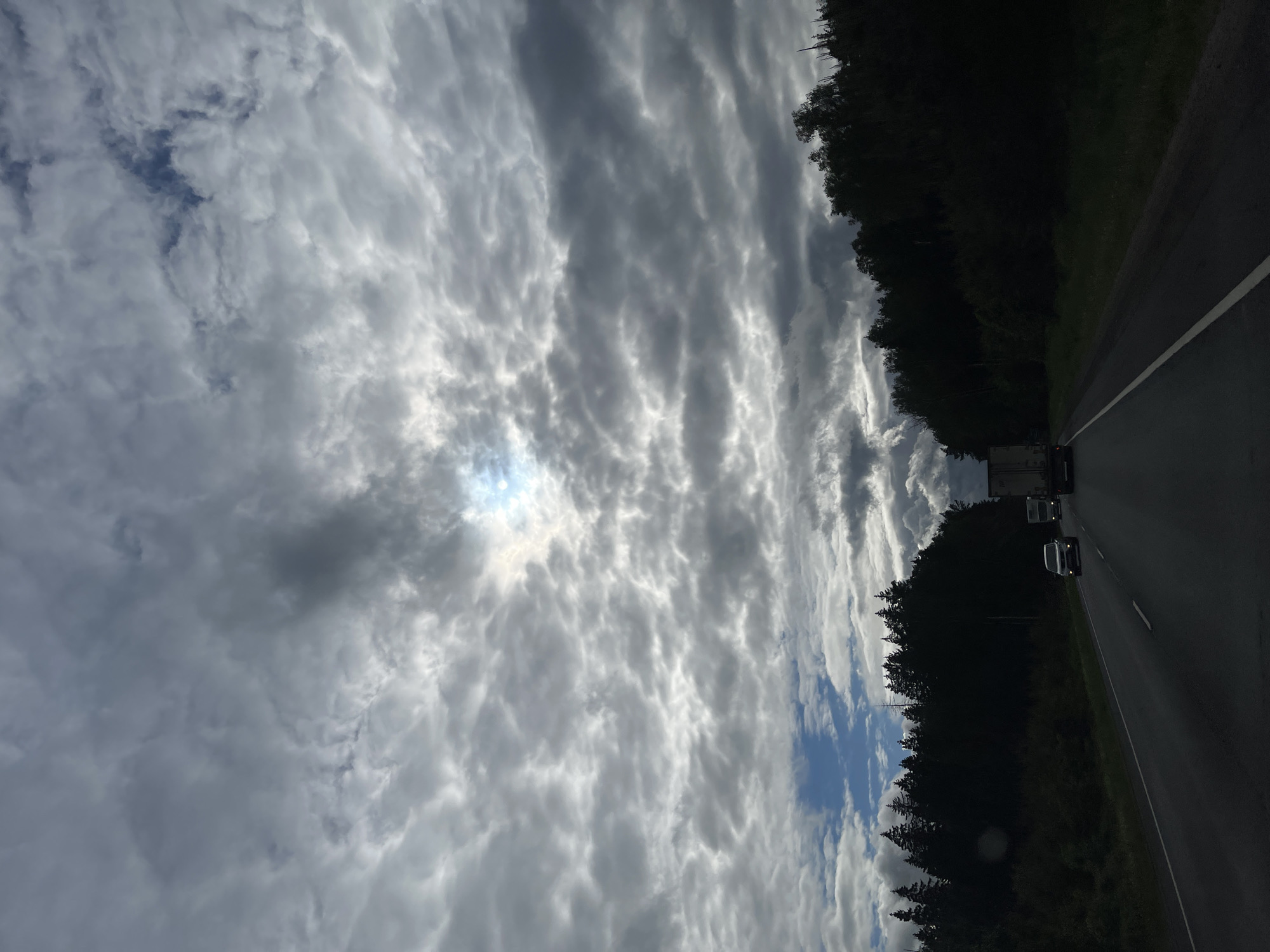 The image shows a road view from a moving vehicle, likely a car, in Ivanovskoye, Russia. The road has two lanes, with a white line separating them. There are at least two vehicles in front, including a truck. Both sides of the road are lined with dense trees. The sky is filled with heavy, textured clouds, with some patches of blue visible. The sun appears to be trying to break through the clouds. The image is oriented vertically, suggesting it may have been taken with a mobile device.
