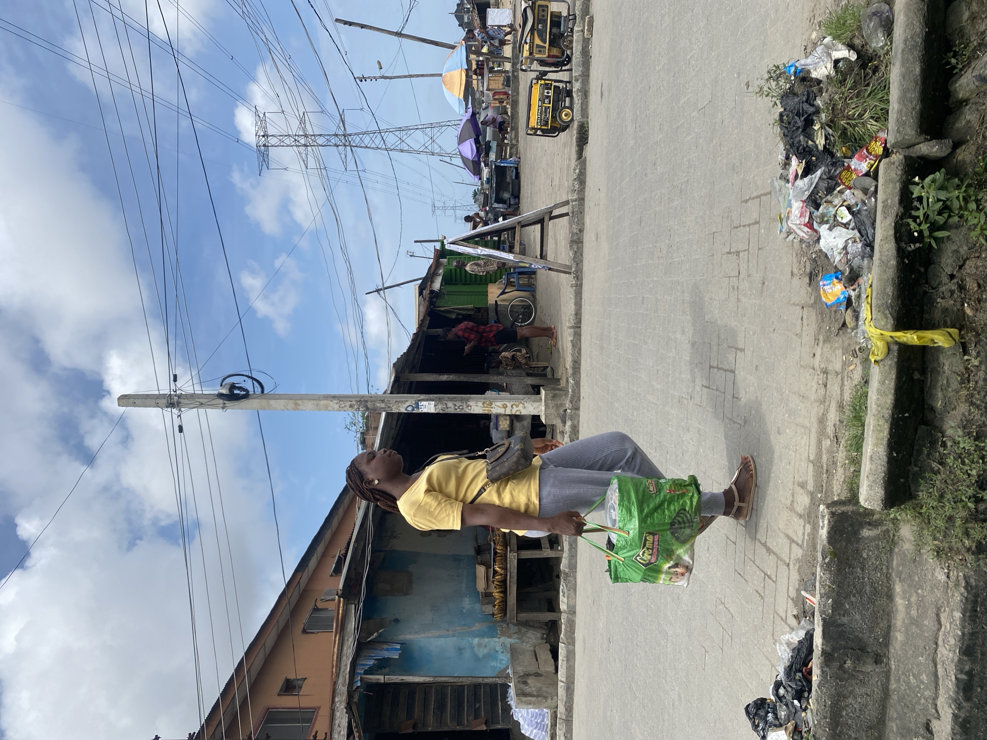 A young Black woman with braided hair walks on a paved sidewalk, carrying a large green shopping bag in her left hand and wearing a yellow t-shirt, gray pants, and sandals. A small crossbody purse is slung over her right shoulder. She is walking past a row of dilapidated market stalls constructed from wood and corrugated metal. The stalls have open fronts displaying goods like bananas and other items. In the background, a large metal electricity pylon and numerous overhead power lines dominate the sky, which is partly cloudy. Two yellow generators are parked on the sidewalk near one of the stalls. A pile of garbage is visible on the edge of the sidewalk in the foreground.