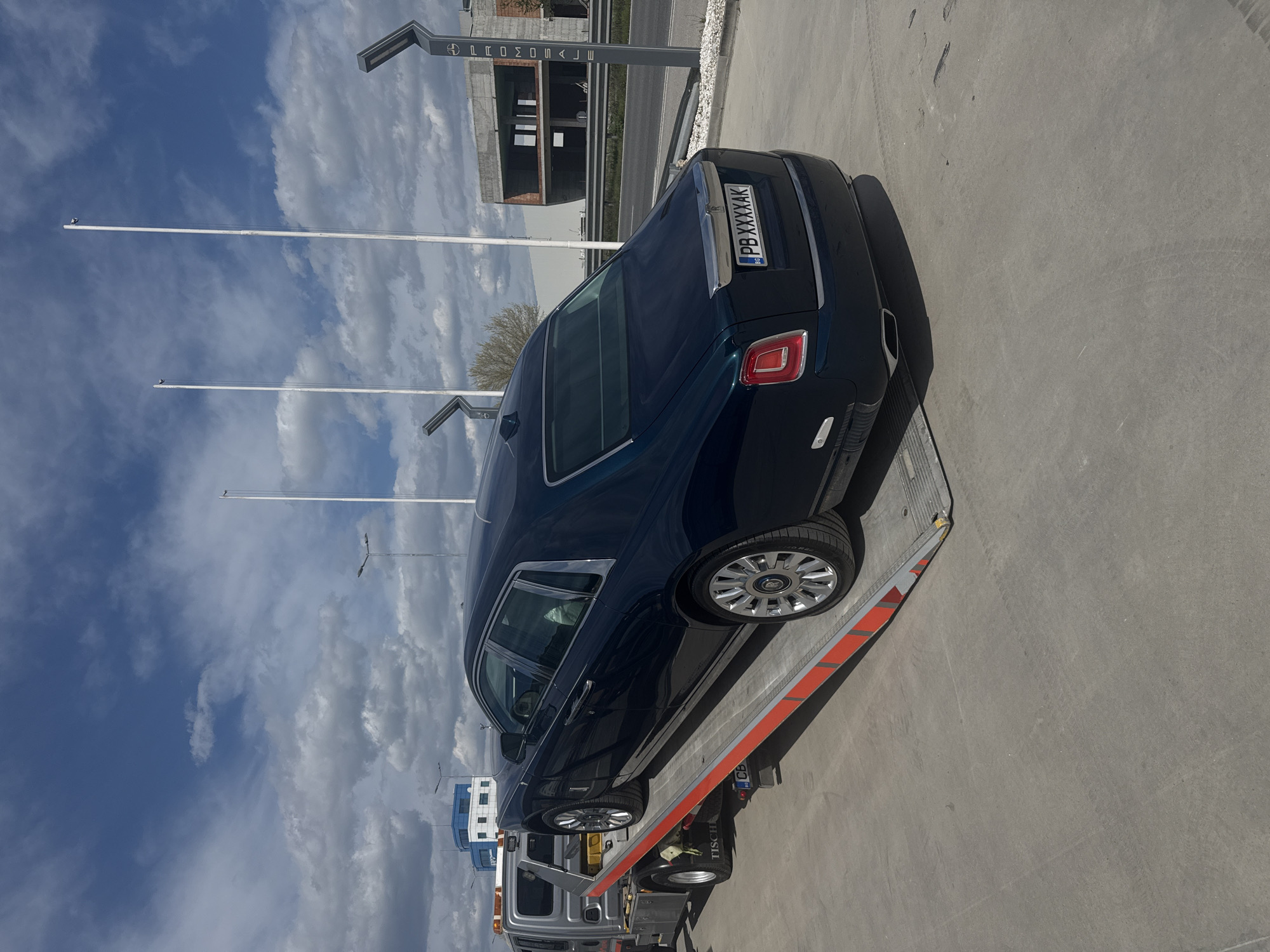 The image depicts a dark-colored Rolls Royce sedan on the bed of a tow truck. The license plate is visible, reading "PB XXXXAK" and indicating a Bulgarian registration. The tow truck is parked on a paved surface. In the background, there are tall poles, a building under construction, and a blue sky with clouds. The image appears to have been taken in Plovdiv, Bulgaria.