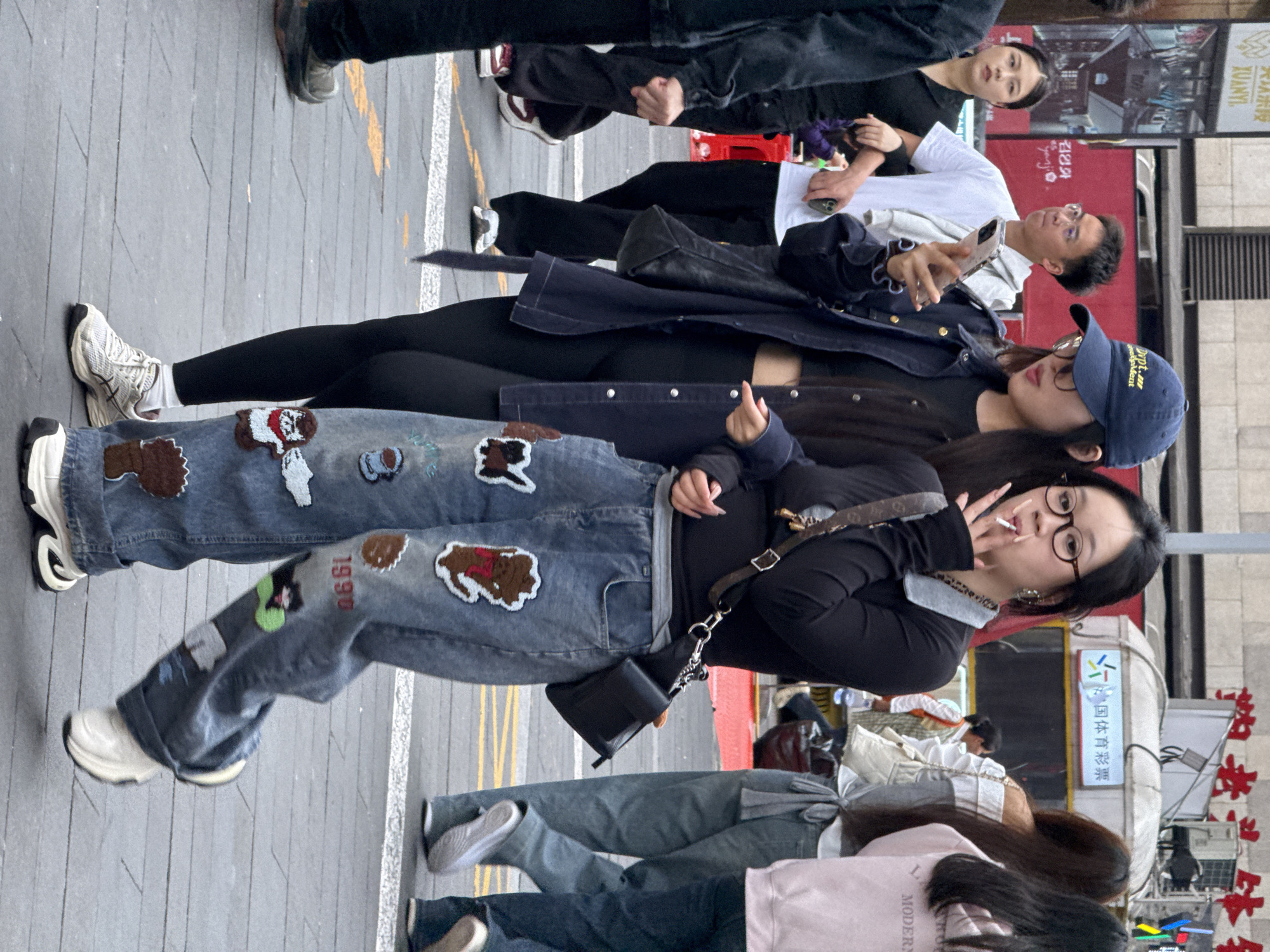 On a paved street in Liangjiang, China, several people are present. One individual with dark hair, wearing glasses, a black top, and jeans with patches, holds a cigarette and faces the camera. Next to her is another person wearing a denim jacket and a dark baseball cap, also facing the camera. Further back, a man with glasses is looking at his phone, while another person stands beside him. The background features red signage with Chinese characters and a booth displaying sports lottery information. Street markings are visible on the pavement.