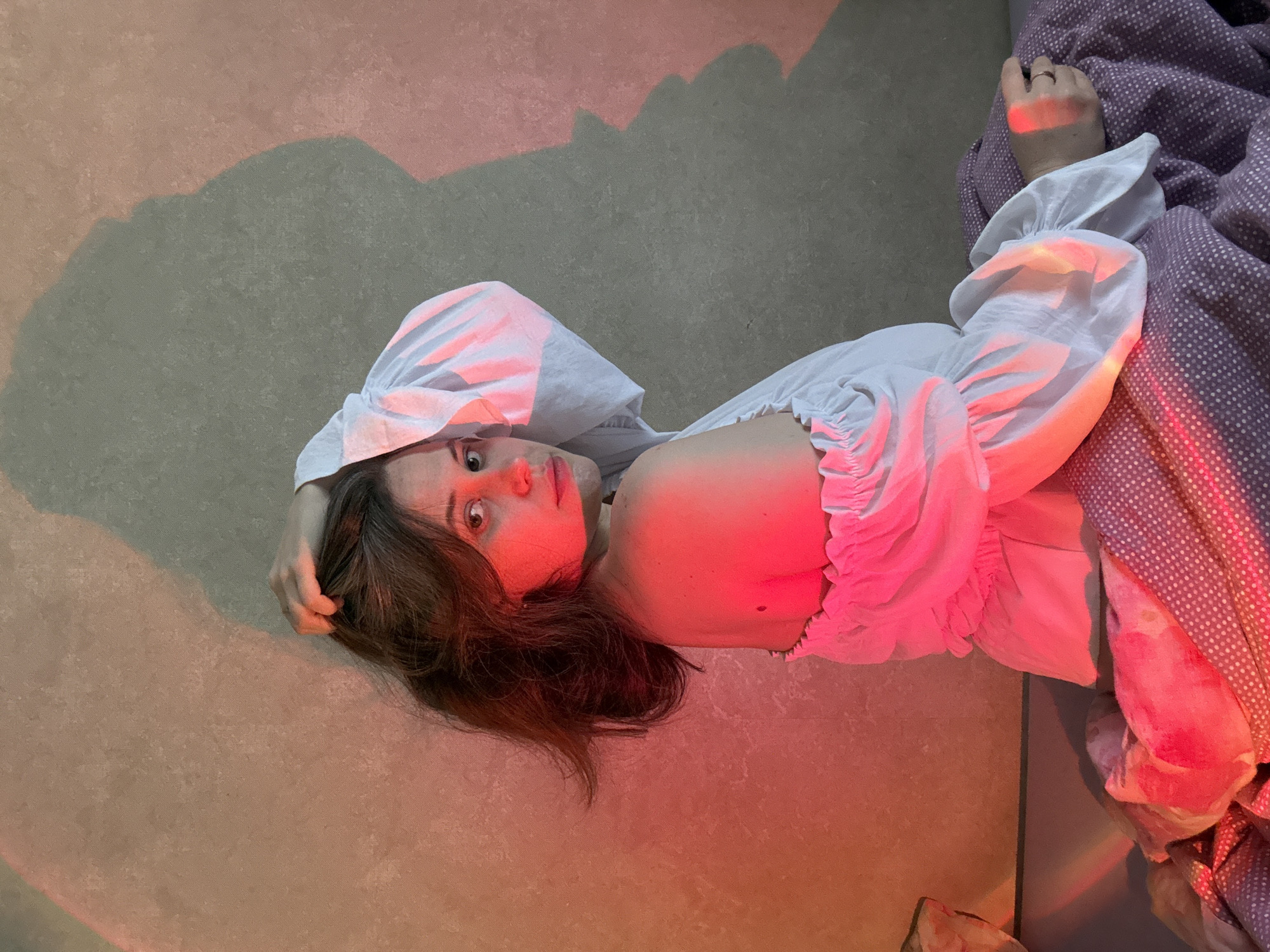 A young woman lies on a gray floor in Chaikovsky, Russia, wearing a white blouse with ruffled sleeves. She is partially covered by a purple polka-dot blanket. A red light illuminates her face and parts of her clothing and blanket. A shadow is cast on the floor next to her.