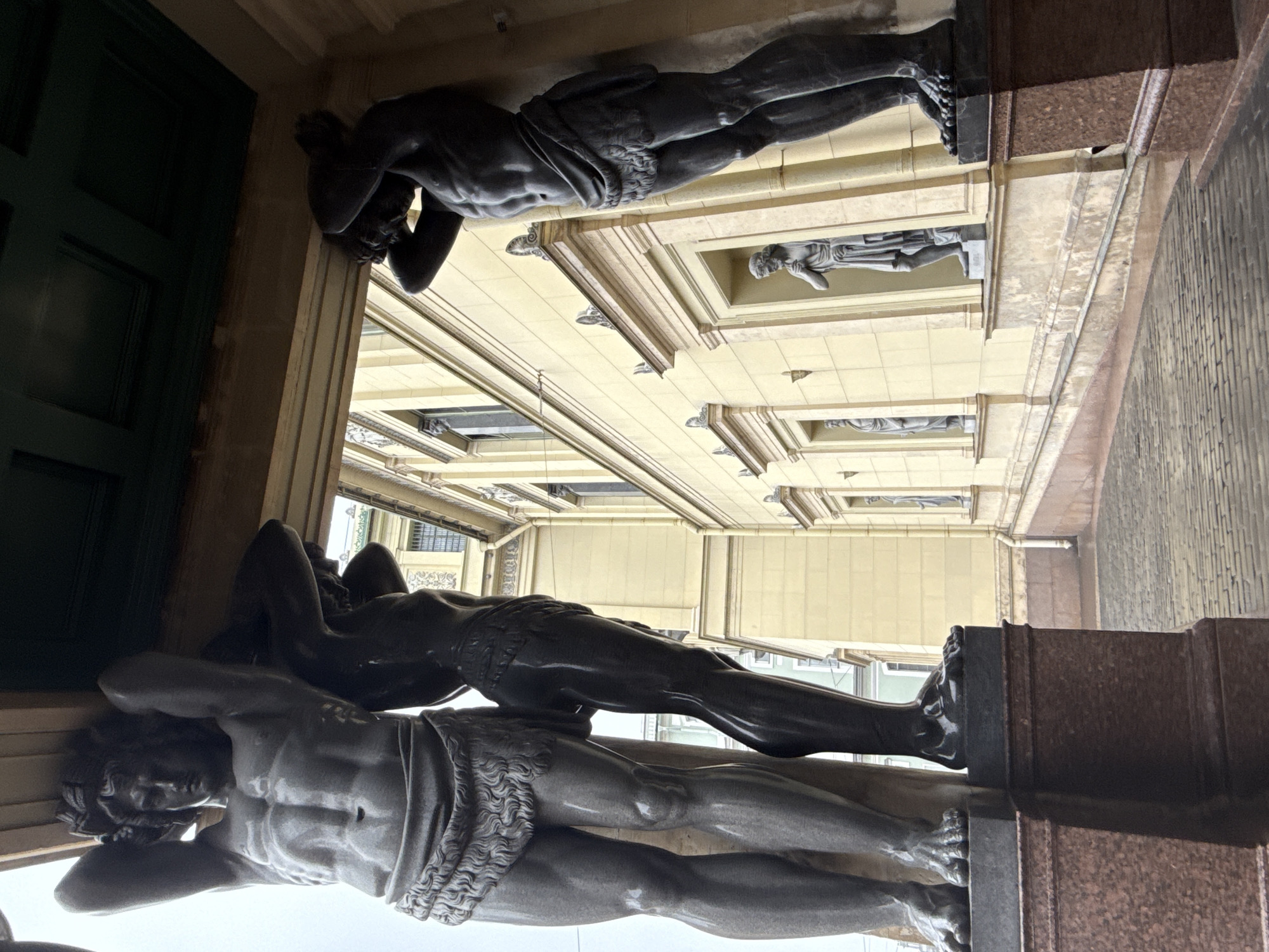 The image shows a view looking upwards at the Hermitage Museum in Saint Petersburg, Russia. The focus is on the architectural details of the building's facade, particularly the sculptures of reclining male figures that appear to be supporting the structure above. The figures are located at the entrance of the museum. There are other sculptures depicting standing people along the façade. The building has a pale yellow facade and intricate architectural details. A dark green door is visible in the background.
