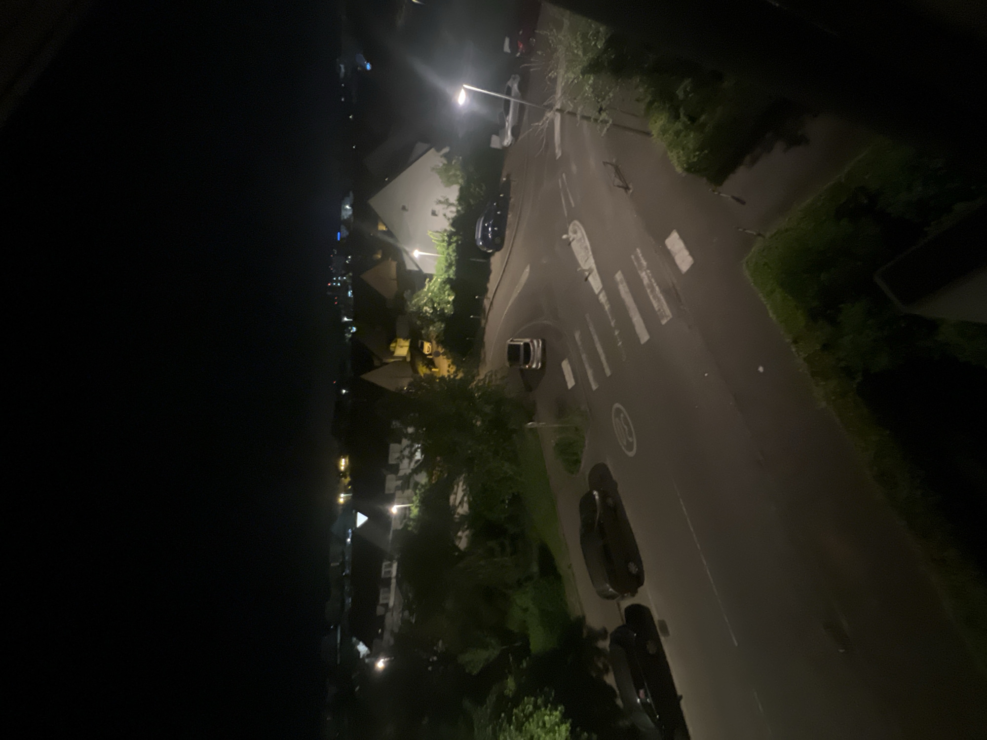 The image shows a nighttime street scene in Strasbourg, France. The perspective is from an elevated position, possibly from a building window. A street with asphalt pavement is visible, marked with a pedestrian crosswalk and a speed limit sign (likely 30 km/h). Several parked cars line the street, and streetlights illuminate the area, casting bright glares. Buildings and trees are visible on either side of the street, creating a suburban environment. In the distance, more lights from buildings and streetlights can be observed, hinting at a broader urban landscape. The left side of the image is predominantly dark, contrasting with the illuminated street scene.