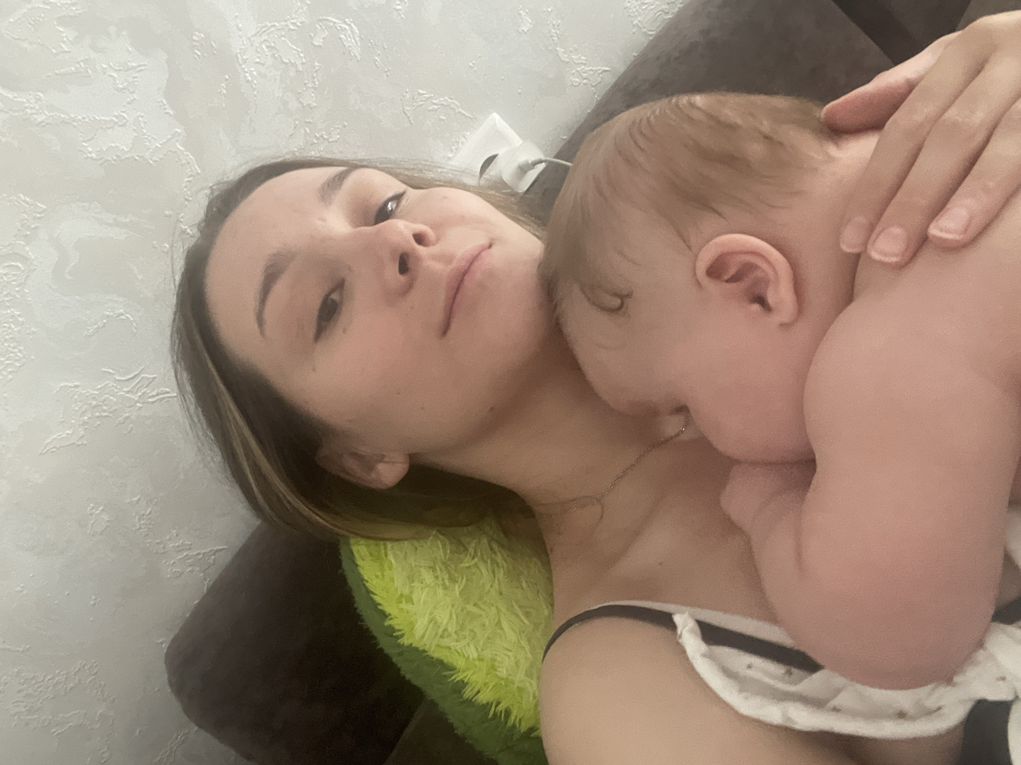 A woman and a baby are lying down, likely on a bed or sofa, in a room. The woman is holding the baby, who appears to be sleeping or resting. A bright green pillow is visible behind them. The wall in the background has a textured surface. There is a white electrical outlet with a cord plugged into it on the wall. The lighting suggests it is indoors, possibly in the late afternoon or evening. The overall scene conveys a sense of intimacy and rest.