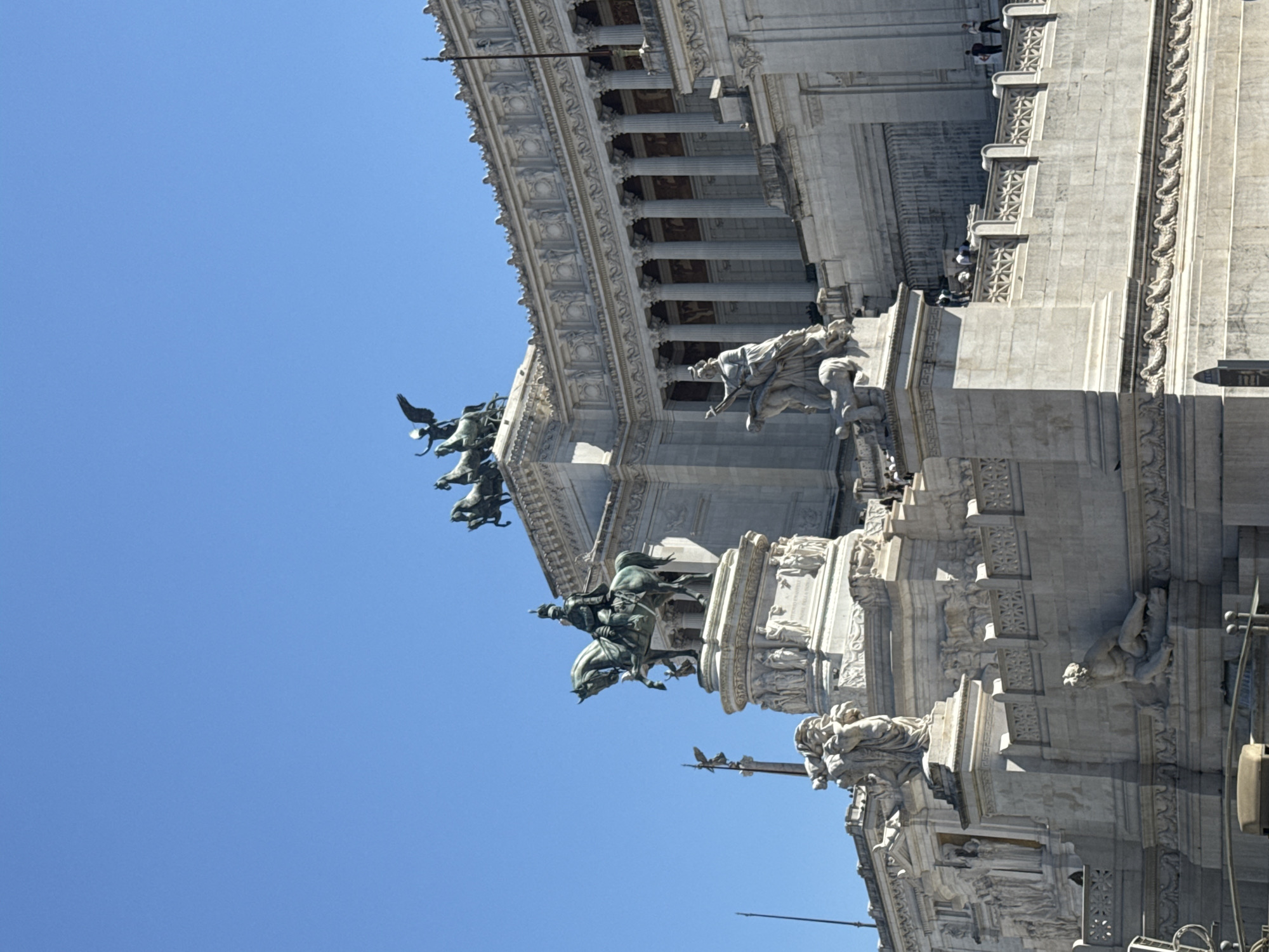The image shows a portion of the Altare della Patria (Altar of the Fatherland), also known as the Monumento Nazionale a Vittorio Emanuele II, in Rome, Italy. The monument is constructed of white marble and features elaborate sculptures, including statues of figures on horseback and winged sculptures. A classical colonnade is also visible. People are walking along the top of the monument. The sky is clear and blue.
