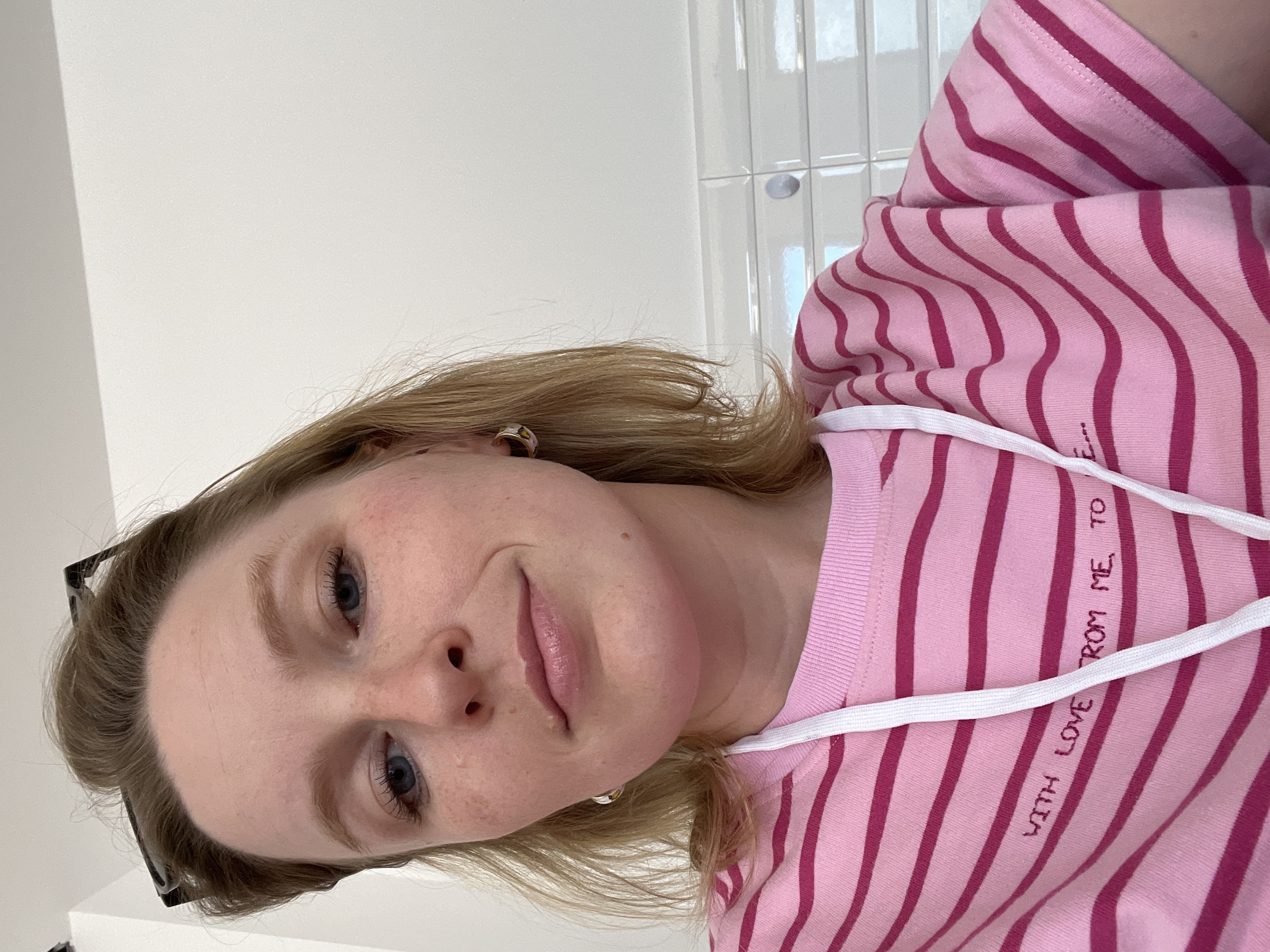 A light-skinned female with blonde hair is pictured in a selfie-style shot. She has blue eyes and a slightly smiling expression. A pair of black glasses are partially visible on her head. The background consists of a plain white wall and a section of white tiled wall. The female is wearing a pink and magenta striped sweatshirt with white drawstrings. Text, partially obscured, is printed on the sweatshirt. The location of the photo is not discernible in the city of Budva, Montenegro.