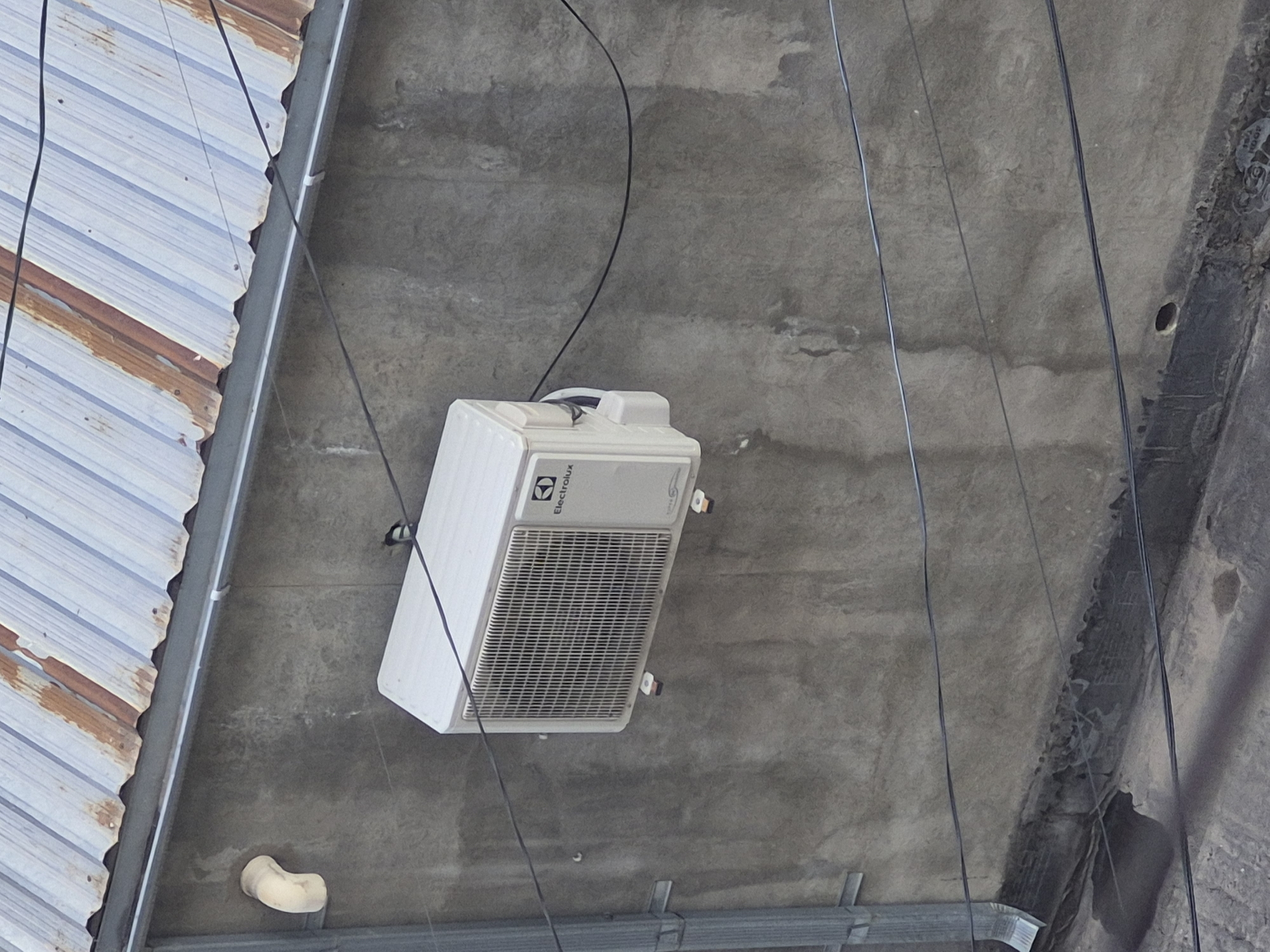 The image provides an upward-angled view of an outdoor utility area, likely part of a building's exterior in Yerevan, Armenia. The central subject is a white Electrolux air conditioning unit, mounted horizontally on a textured, grey concrete surface. The unit features a prominent grille on its underside and the "Electrolux" brand name is visible on its side.
To the left, a corrugated metal roof, showing signs of wear and rust, borders the scene. A grey metal gutter runs parallel to the roof's edge. Multiple black and dark grey wires or cables traverse the concrete background, connecting various points within the frame.
The concrete surface exhibits variations in texture and discoloration, suggesting age or exposure to the elements. A light-colored, L-shaped pipe or drain elbow is affixed to the concrete towards the bottom left. On the right side, a dark, rough-surfaced structural element runs vertically, with a small, circular hole visible in the adjacent concrete.
The bright, even lighting indicates it is daytime. No people or activity are present, and the scene is static, highlighting functional building components and infrastructure. The overall impression is that of an industrial or service area of a building.