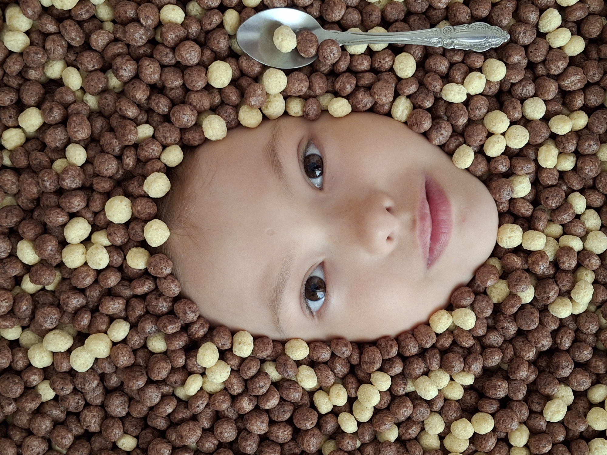 A child's face is surrounded by a pile of round, brown and light-yellow cereal balls. A silver spoon containing a single light-yellow cereal ball rests on top of the child's head. The background consists entirely of the cereal balls, with no other objects visible. The location is unknown and there is no information in the image that relates to Chaikovsky, Russia.