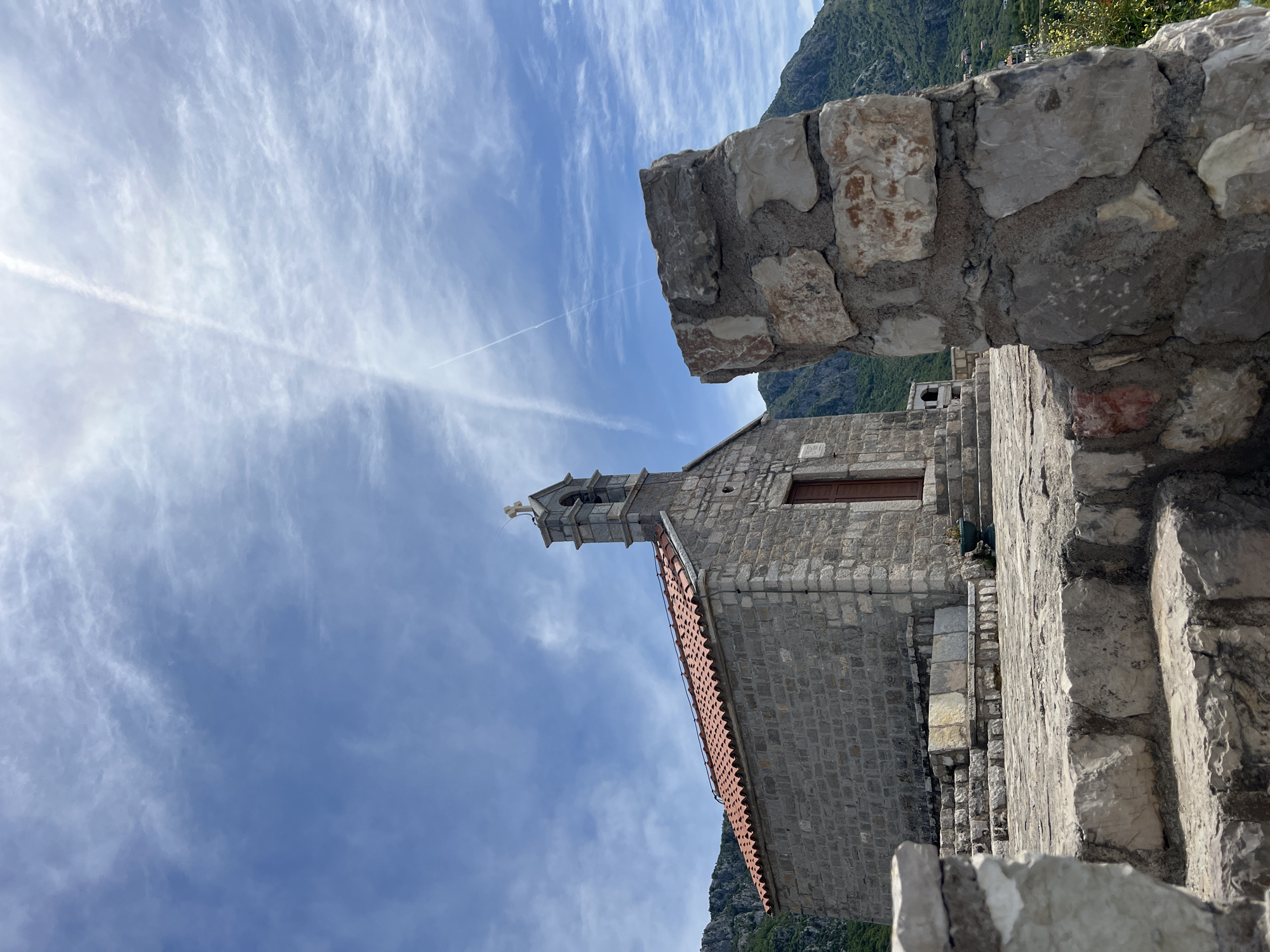 Captured from a low angle, the image presents a stone building with a steeple and cross against a sky marked by wispy clouds and a contrail. The building, likely a chapel or church, features a stone façade with a contrasting orange tiled roof, and is viewed from a vantage point framed by rough stone walls and steps. This building is located in Sveti Stefan, Montenegro.