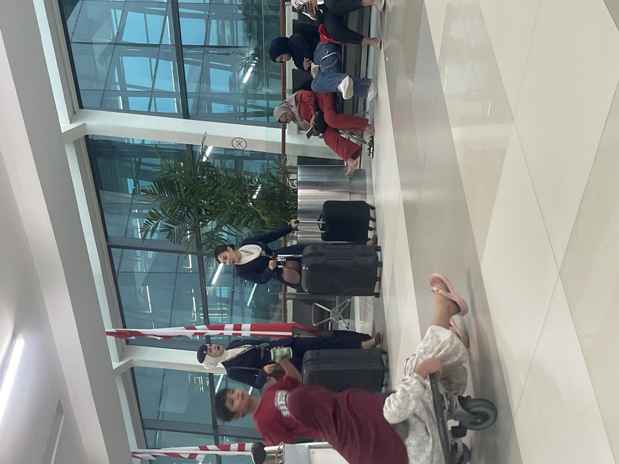 The image depicts a scene at an airport in Tangerang, Indonesia, likely within the departure or arrival hall. In the foreground, a child is being pulled on a luggage trolley, wearing a red top and patterned trousers, with pink flip-flops visible. To the left of the child, an individual wearing a maroon headscarf and matching trousers sits facing away from the camera.
In the mid-ground, two women in flight attendant uniforms stand near large black suitcases. The woman on the left, wearing a white hijab, holds a green can. The woman on the right, with a scarf around her neck, leans against a suitcase. In the background, several people are seated on a bench, appearing to wait. One woman, wearing a red outfit and hijab, is looking at her phone, while another person in a dark blue hijab and jeans is also seated. The airport environment is characterized by large windows reflecting the exterior, flags, and overhead lighting. A potted plant adds a touch of greenery to the scene.