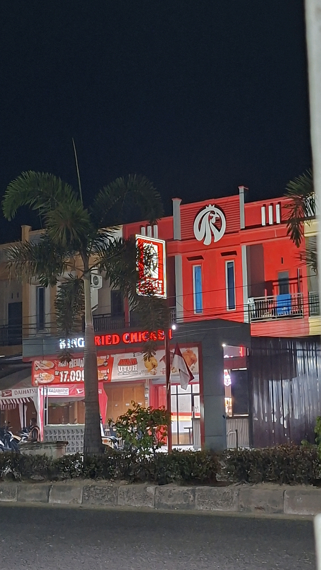 The image depicts a street scene at night in Lhokseumawe, Indonesia. A brightly lit red building with a prominent white bird logo at the top is the main focus. This building appears to be a restaurant, likely a fried chicken establishment, as indicated by the signage "KING FRIED CHICKEN". Below the main building, there are smaller signs advertising food items, including one with "AYAM UTUH" (whole chicken) and prices like "17.00!". There are also signs for "DAIHATSU" which suggests a nearby car dealership or service center. Palm trees are visible in the foreground, and the street in front of the building has a sidewalk with a low concrete barrier and some low-lying green bushes. The overall atmosphere is one of commercial activity at night.