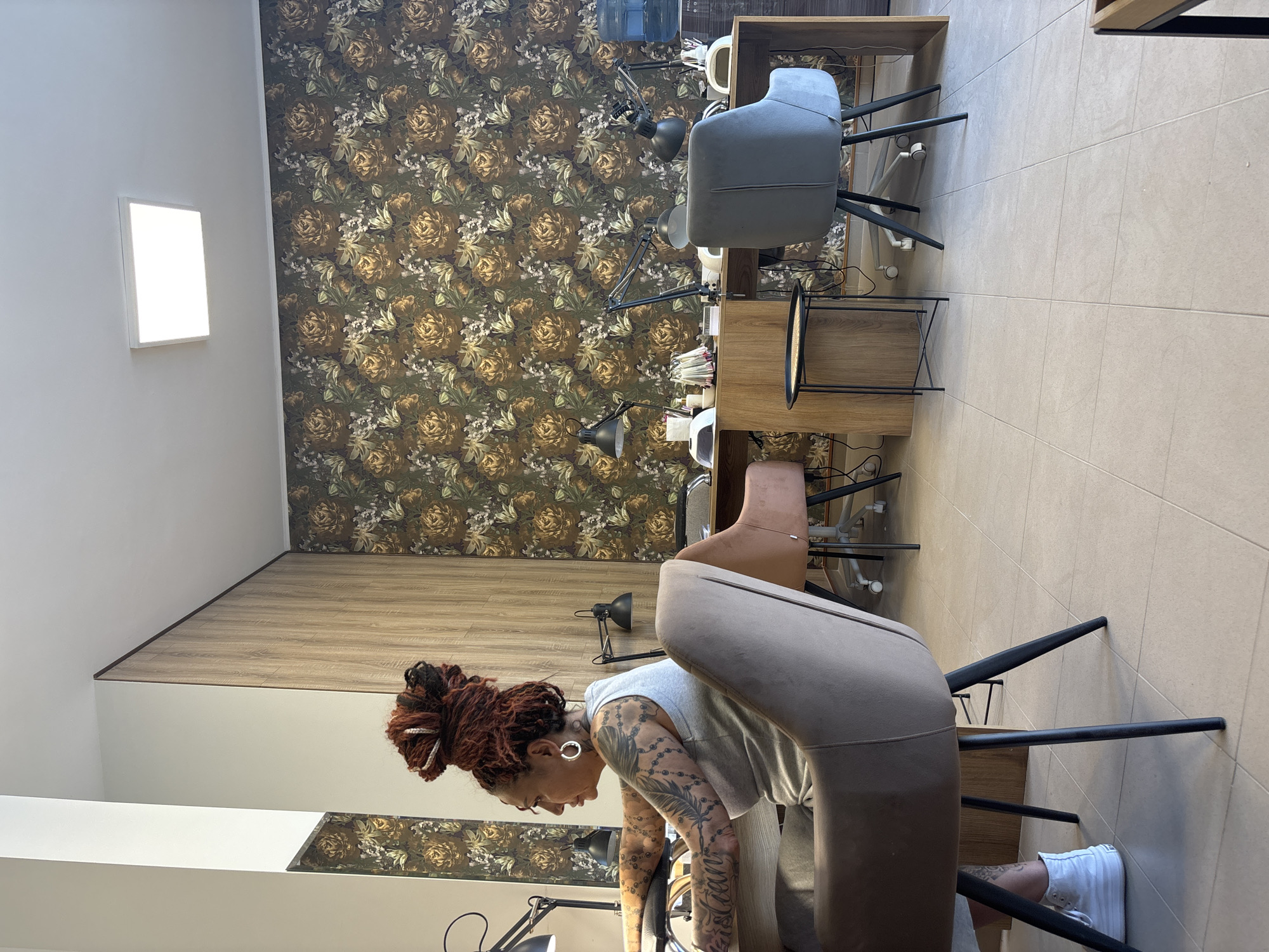 The image depicts the interior of a nail salon, likely located in Plovdiv, Bulgaria. A woman with reddish-brown dreadlocks styled in a top bun is seated at a workstation, facing to the left. She is wearing a grey tank top and grey pants, and white sneakers. Her arms and shoulders are covered in tattoos. She appears to be working on something at the workstation, though her hands are not fully visible.
The workstation is equipped with multiple nail lamps and various tools for nail services. In the background, two other workstations are visible, each with similar equipment. The walls are adorned with a floral wallpaper featuring large, stylized yellow and brown roses against a dark green background. A large mirror is partially visible on the left wall, reflecting the floral wallpaper and a light-colored wall. The flooring is tiled in a light beige color. Two chairs are positioned at the workstations; one is a light brown upholstered chair, and the other is a dark grey upholstered chair with wheels. A small, round side table with a woven top is placed between the chairs. The overall atmosphere suggests a professional beauty salon.