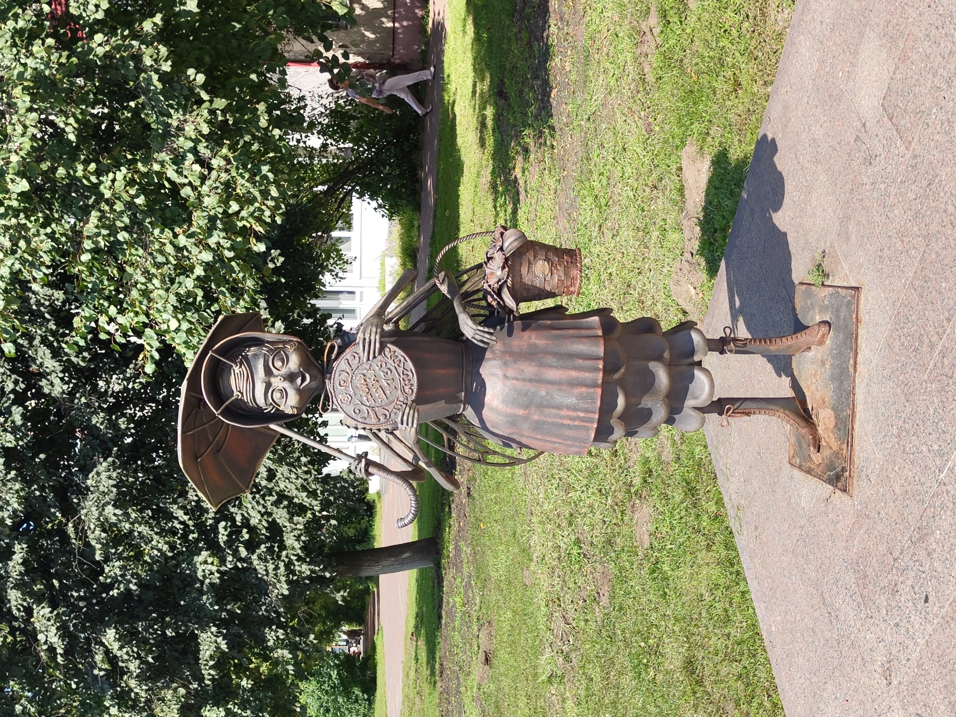 This is a medium shot, eye-level, outdoor photograph of a metal sculpture of a stylized bee-like character. The sculpture is standing on a granite base in a grassy park area. The character is anthropomorphic, with large, expressive eyes, a small mouth, and antennae. It is wearing a ruffled skirt, boots, and a hat resembling an umbrella. In its left hand, it holds a round, decorative plate, and in its right hand, it carries a woven basket filled with what appears to be flowers or leaves. The sculpture is cast from a dark metal, possibly bronze or iron, and exhibits a weathered patina.
Behind the sculpture, a paved walkway runs diagonally from the left to the right of the frame. A person, casually dressed in light-colored pants and a patterned shirt, is walking away from the viewer on this walkway. Lush green trees with dense foliage form a backdrop to the scene, with sunlight filtering through the leaves and casting shadows on the ground. In the distance, a white building with windows is partially visible through the trees. The overall environment suggests a public park or garden in a temperate climate during daytime, under a clear sky.
This sculpture is located in the city of Ramenskoye, Russia. Specifically, it is situated in the central park of culture and leisure of Ramenskoye, a popular public space. The sculpture is known locally as "The Bee" or "Little Bee" and is a beloved landmark in the city, often associated with the historical importance of beekeeping in the region.