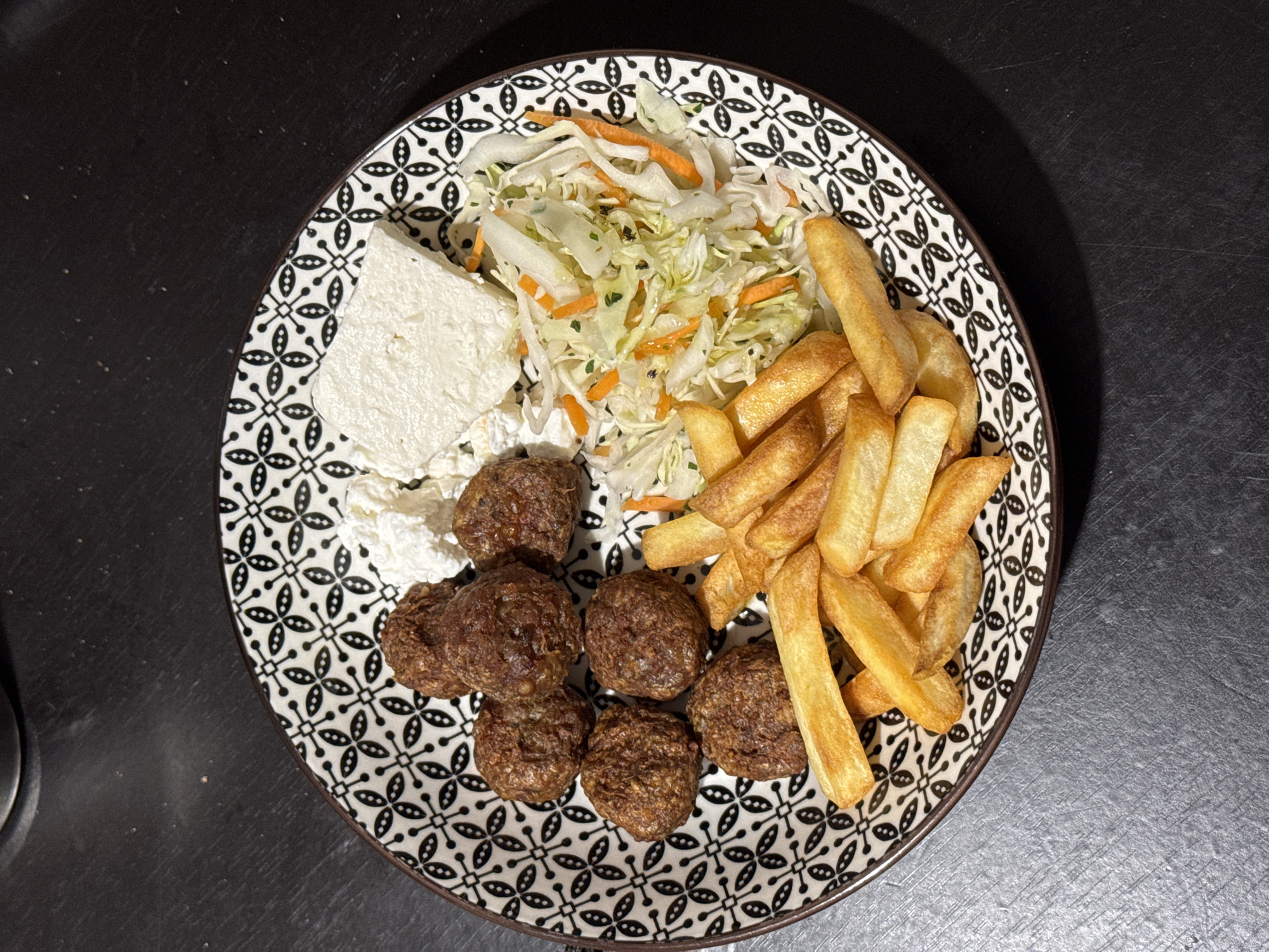 An overhead, close-up shot shows a dinner plate filled with food. The plate has a black and white geometric pattern around the rim. In the center of the plate, there are seven meatballs, two pieces of white cheese, a pile of french fries, and a small salad of shredded cabbage and carrots. The food is arranged on a black surface that appears to be a table.
