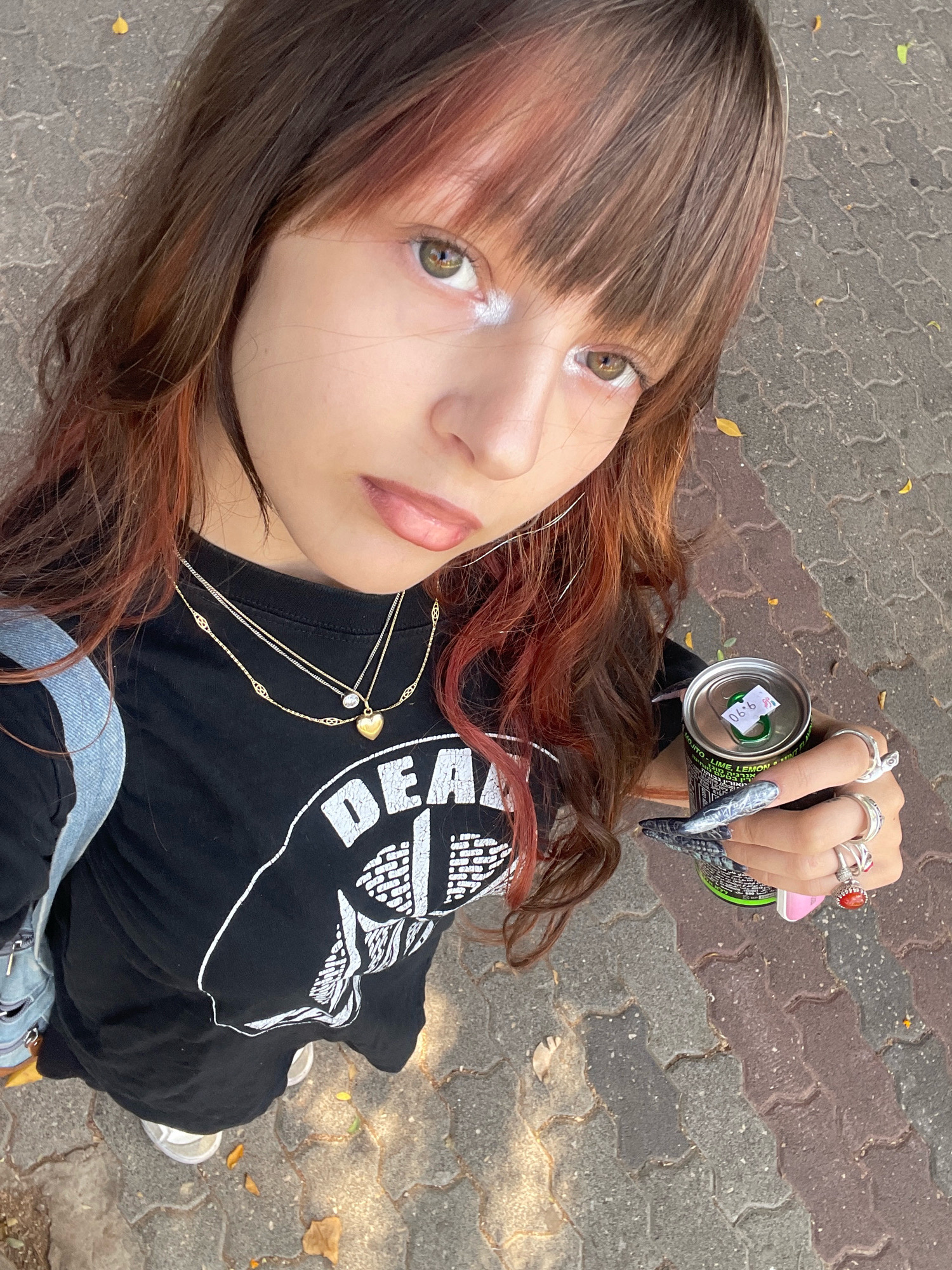 The image is a close-up, high-angle selfie taken outdoors on a paved sidewalk in Raanana, Israel. The subject is a young person with long, brown hair streaked with reddish tones and bangs. They are wearing a black t-shirt with a white graphic design, multiple necklaces, and several rings on their fingers. The person is holding a green can of energy drink in their right hand and has distinctive, elongated, patterned fingernails. The background consists of the textured pavement of the sidewalk, with a few fallen leaves visible. The lighting suggests it is daytime, with sunlight casting shadows on the ground.