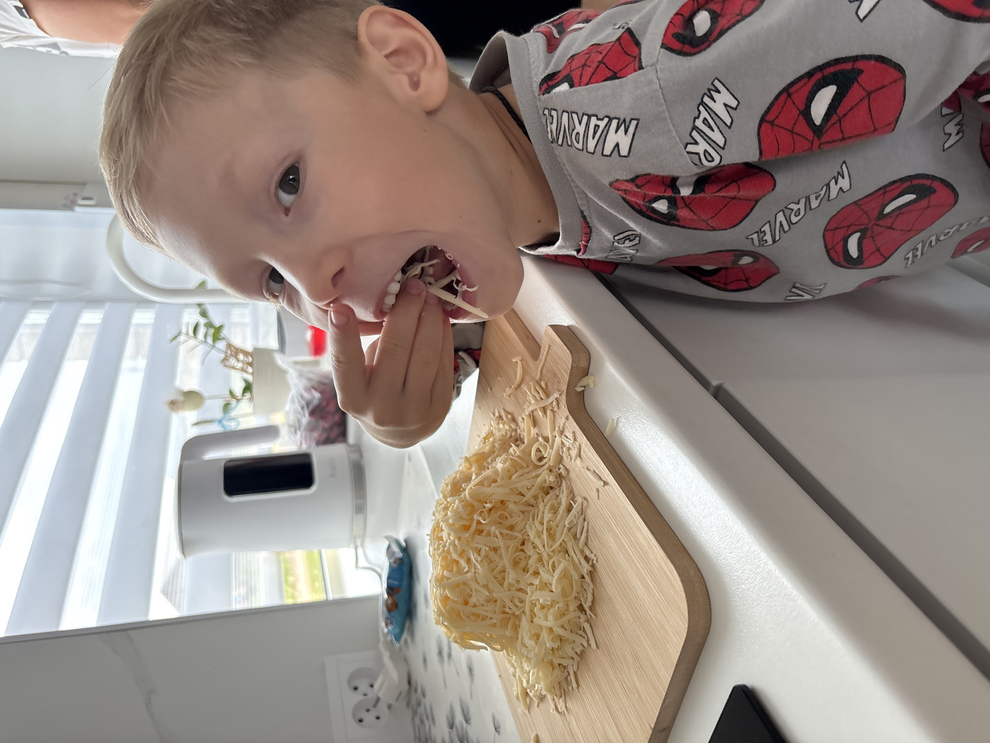In this close-up, portrait-oriented image, a young Caucasian boy with blond hair is the central subject, looking directly at the camera with a playful expression. He is in the process of eating shredded cheese, with some of it in his mouth and his finger reaching towards it. He is wearing a gray t-shirt adorned with a repeating pattern of Spider-Man masks and the Marvel logo.
The setting appears to be a kitchen, indicated by the presence of a white electric kettle with a black display on the counter to the left, and a wooden cutting board with a pile of shredded cheese on it in the foreground. The countertop is white and appears to be made of a solid surface material. In the background, a window with white blinds is visible, suggesting it is daytime. The overall lighting is bright and natural.
The boy's open mouth and curious gaze convey a sense of playful mischief. The image is shot at a slightly tilted angle, adding to the candid and dynamic feel of the moment. There is no discernible weather information. The visible text on the boy's shirt includes "MARVEL" repeated in a stylized font. The overall mood is lighthearted and captures a child enjoying a snack.