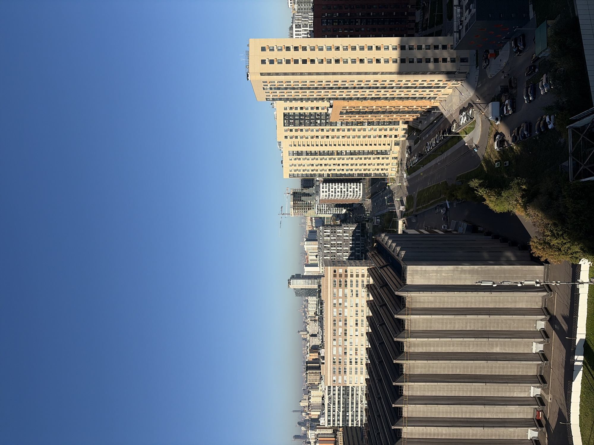 This is an aerial view of a city, likely Moscow, Russia, on a clear day. The foreground is dominated by a large, grey, industrial-style building with a sloped, ribbed roof. Its facade is vertical concrete with regularly spaced openings at the base. To the right of this building, a street runs diagonally away from the viewer, lined with parked cars. Beyond the street, several multi-story residential buildings are visible, varying in color and architectural style, including beige and darker brick buildings with numerous windows. In the background, a dense cityscape extends to the horizon, with taller skyscrapers and construction cranes indicating ongoing development. The sky is a vibrant, cloudless blue.