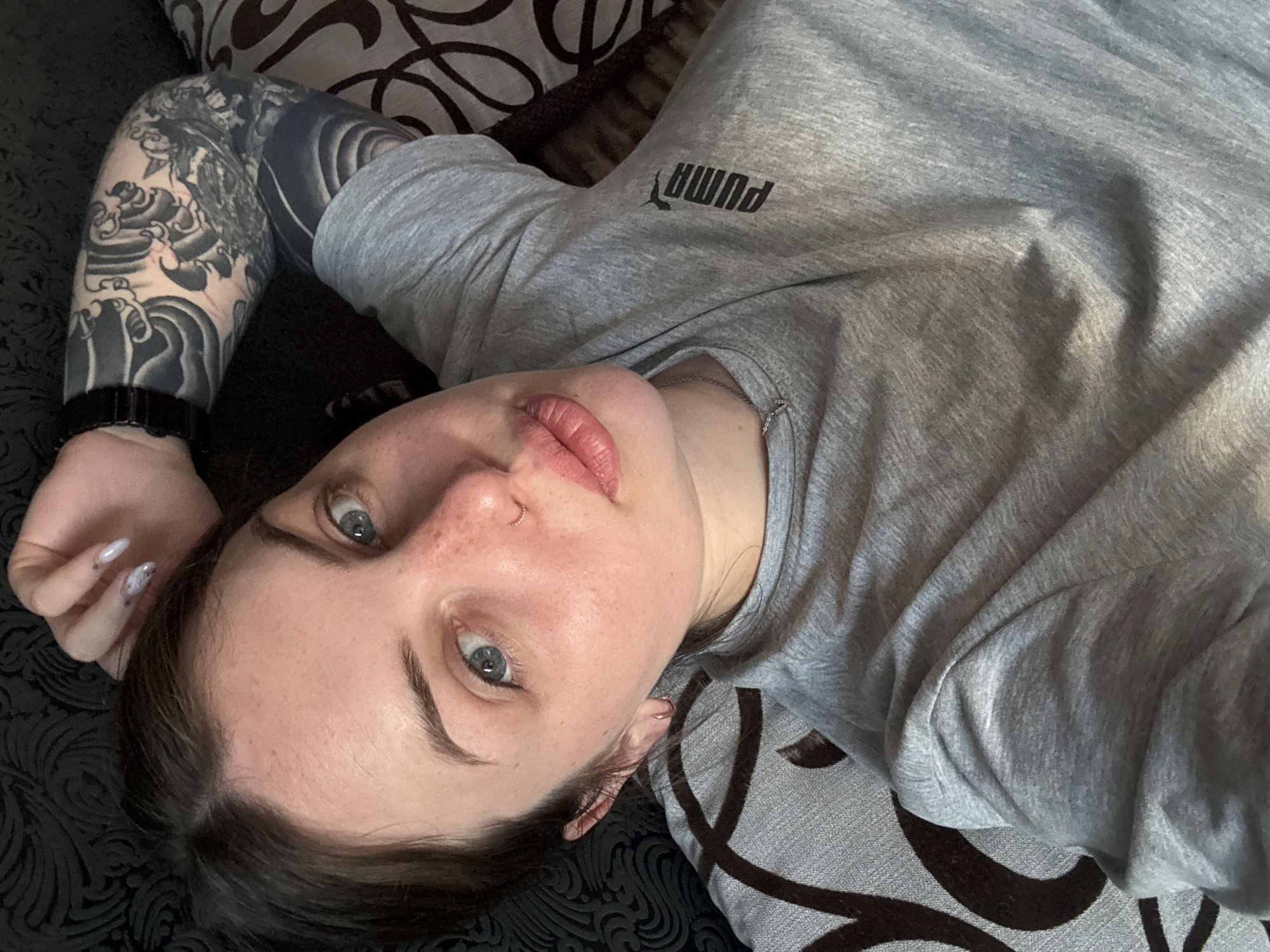 A young woman is depicted lying down, viewed from a slightly elevated angle, looking directly into the camera. She has light skin with freckles scattered across her nose and cheeks, blue-grey eyes, and well-defined dark eyebrows. Her lips are full and slightly parted. She wears a small gold hoop nose ring and a matching earring. Her dark brown hair is visible around her face.
She is dressed in a light grey short-sleeved t-shirt with the "PUMA" logo, printed in black, visible on her chest (appearing reversed). A thin silver chain necklace is also visible. Her left arm is heavily tattooed with a black and grey Japanese-style sleeve featuring detailed waves and traditional motifs; a dark wristwatch is worn on this wrist. Her fingernails are long, pointed, and painted a light, subtly patterned color.
The woman is resting on a surface covered with a dark fabric featuring a light, abstract pattern, likely a sofa or bed. The lighting suggests a bright, natural indoor setting during the daytime. The overall impression is casual and relaxed. The provided location context is Chornomorskoe, Ukraine.