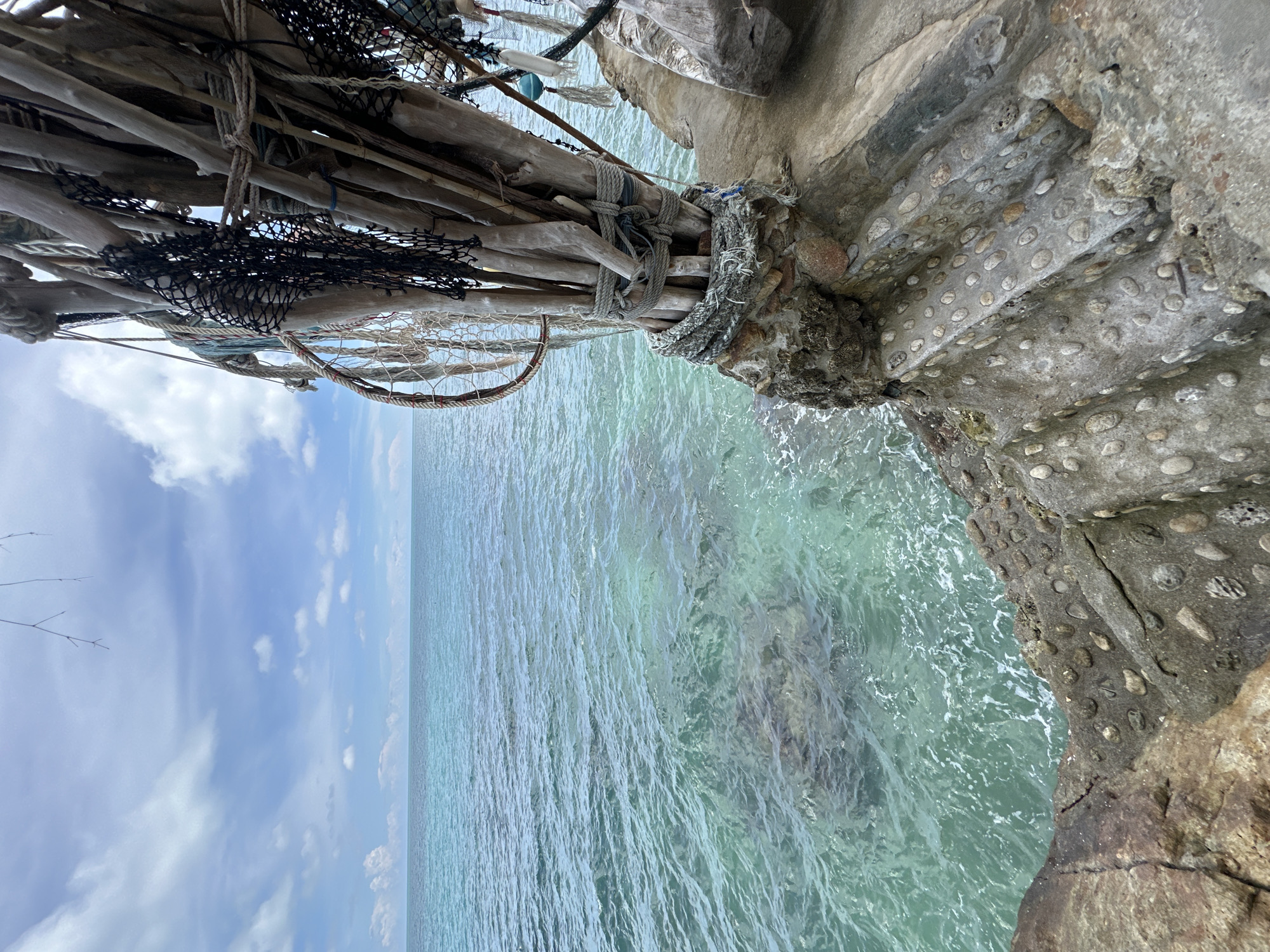 This is a medium-shot, eye-level photograph of a rustic, outdoor structure at the edge of the ocean. The structure is made of weathered driftwood, ropes, and fishing nets, tied together and anchored to a rocky foundation that incorporates a staircase embedded with smooth, rounded pebbles. The staircase leads down to the water's edge. To the left of the structure, the clear, turquoise ocean stretches out to the horizon under a blue sky with scattered white clouds. The water is calm with gentle ripples, and the seabed is visible in the shallows. The scene is located in ตำบลเกาะพะงัน, Thailand.