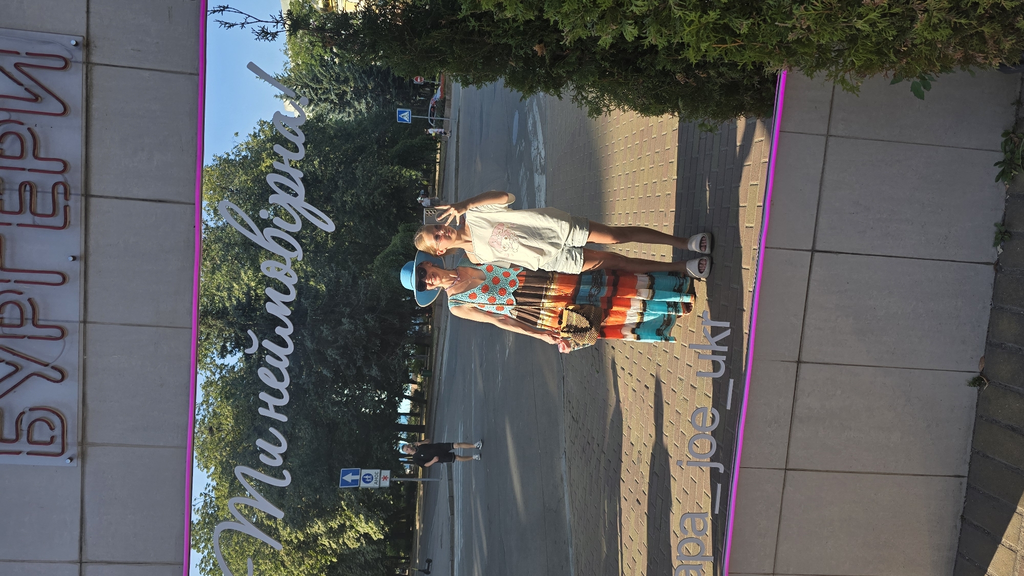In Zhytomyr, Ukraine, a reflective surface mirrors a bright, sunny day, with lush green trees forming a backdrop. Above, the word "БУРГЕРИ" (Burgers) is displayed in neon lettering. Below this, a cursive white inscription reads, "Ти неймовірна!" (You are incredible!). In the foreground of the reflection, two females are captured in a selfie. An older woman, wearing a wide-brimmed blue hat and a colorful, multi-patterned maxi dress with a crochet top adorned with orange circles, stands on the left. She is holding a woven straw purse. To her right stands a younger female, possibly her daughter, wearing a light-colored t-shirt with a cartoon graphic and grey denim shorts. She is also wearing white sandals and sunglasses. The pavement below them is paved with bricks. Along the bottom edge of the reflective surface, a white inscription reads "apa__joe__ukr". A bright pink LED strip runs along the bottom of the reflective panel. In the background of the reflection, a man in a black t-shirt and shorts walks across a street, and a traffic sign is visible.