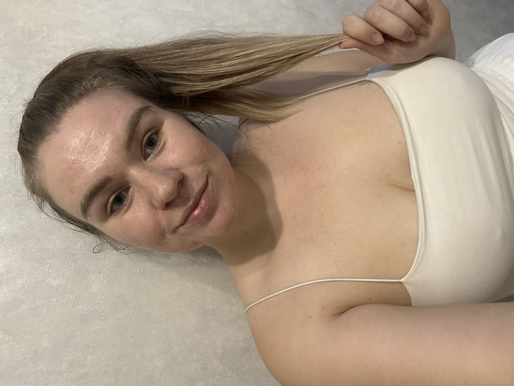 A fair-skinned woman with blonde hair lies on a light-colored surface. She is wearing a white camisole and holding a strand of her hair in her right hand. Her head is tilted toward the camera. The background is a uniform light color. The location is unknown and not discernable from the image.