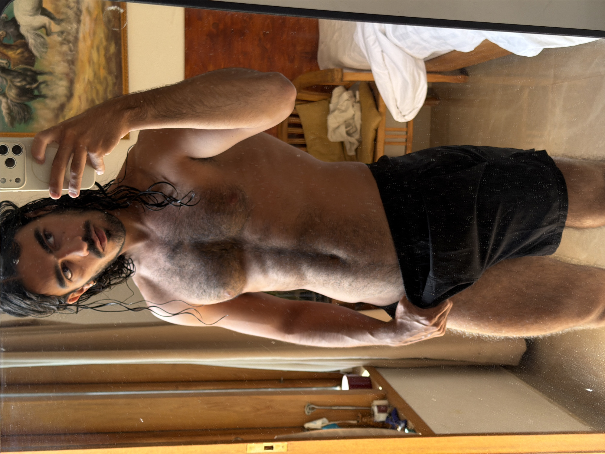 A shirtless man with dark hair, a mustache, and dark shorts is taking a selfie in a mirror. His face is slightly blurred. The reflection in the mirror shows a bed with white sheets and pillows, a painting on the wall, and a wooden structure. Small objects are visible on the lower part of the frame, in the bottom of the mirror's reflection. The exact location in North Kuta, Indonesia is not possible to determine from the image alone.