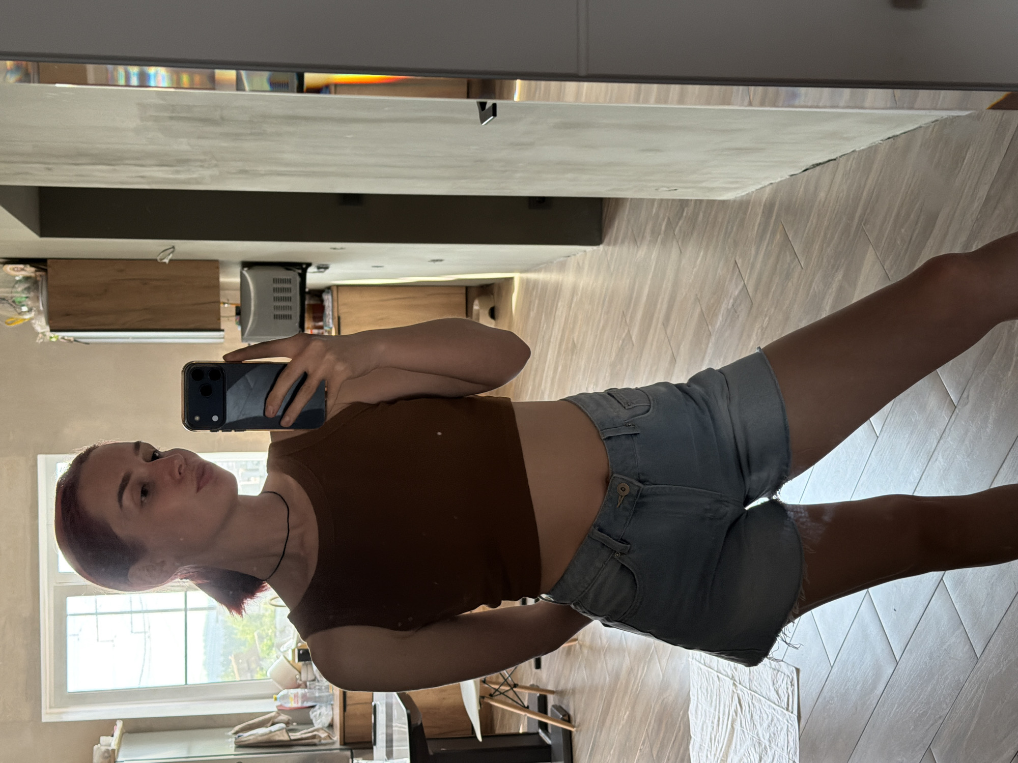 In an indoor setting, likely a residential kitchen or dining area in Chaikovsky, Russia, a young woman with reddish-brown hair tied back in a low ponytail is taking a mirror selfie. She is wearing a brown sleeveless crop top and light-wash denim shorts with a frayed hem. She is holding a dark-colored smartphone in her right hand, angled to capture her reflection.
The background reveals a domestic environment. To the left, a large window offers a view of greenery and possibly hills, suggesting a location with natural surroundings. A dining table and chair are partially visible, as are kitchen cabinets and appliances. The floor is tiled in a wood-grain pattern. To the right, a large mirror reflects the scene, and a doorway with a dark door handle is also visible. The lighting appears to be natural, coming from the window.